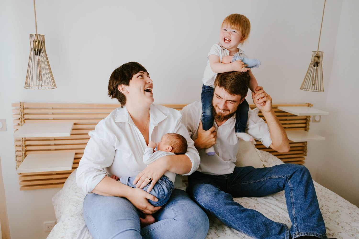 Happy family with newborn baby and toddler on dad's shoulders laughing together on a bed at home.