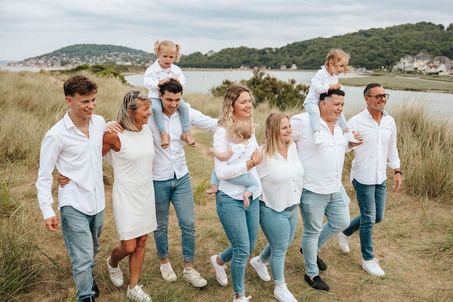 A multigenerational family in white shirts and jeans walks cheerfully through coastal grasslands together.