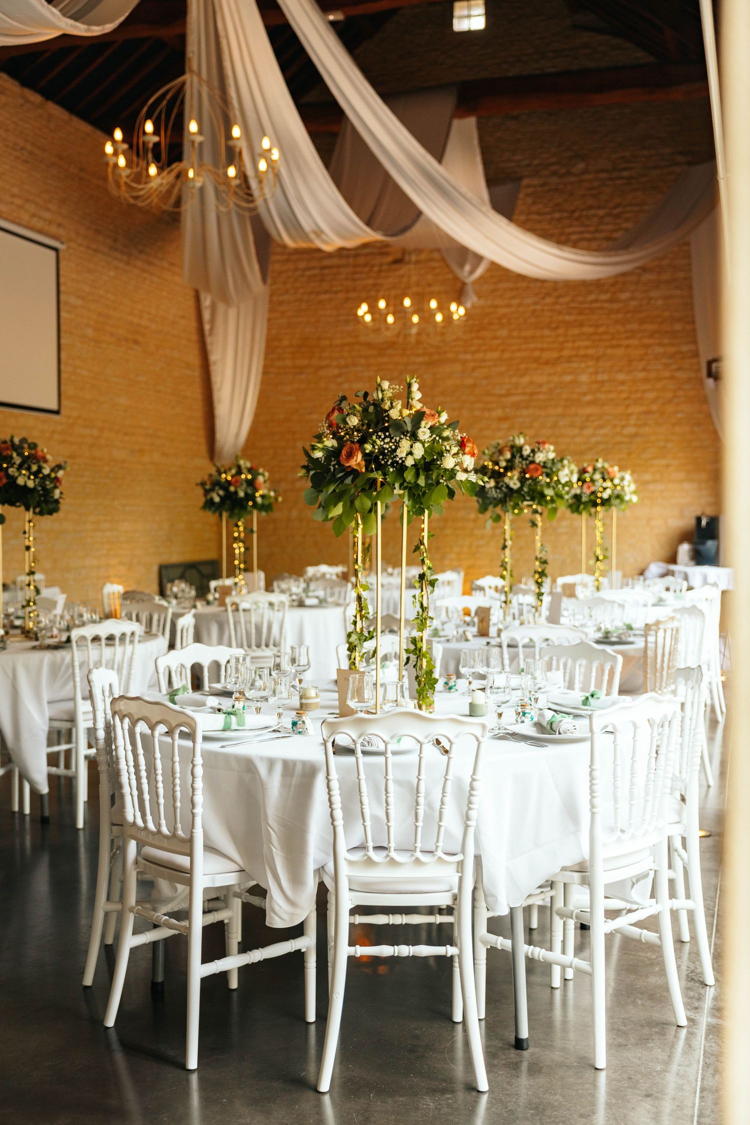 Elegant wedding reception hall with white Chiavari chairs, tall floral centerpieces, white draped ceiling, and gold chandeliers.