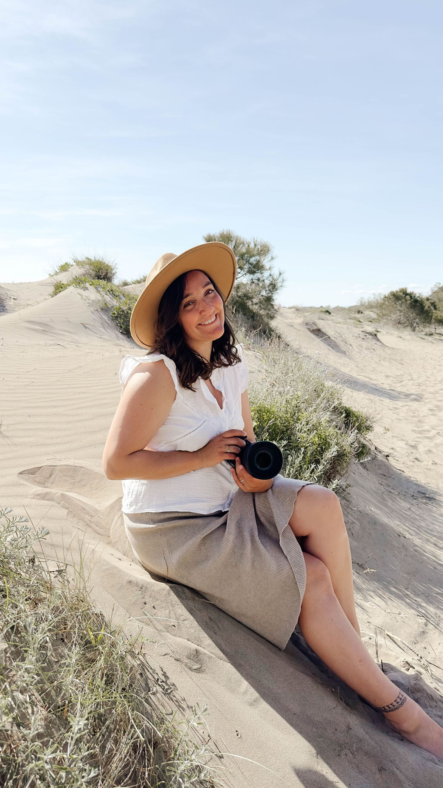Smiling female photographer in hat and white top sitting on sand dunes holding a camera outdoors.