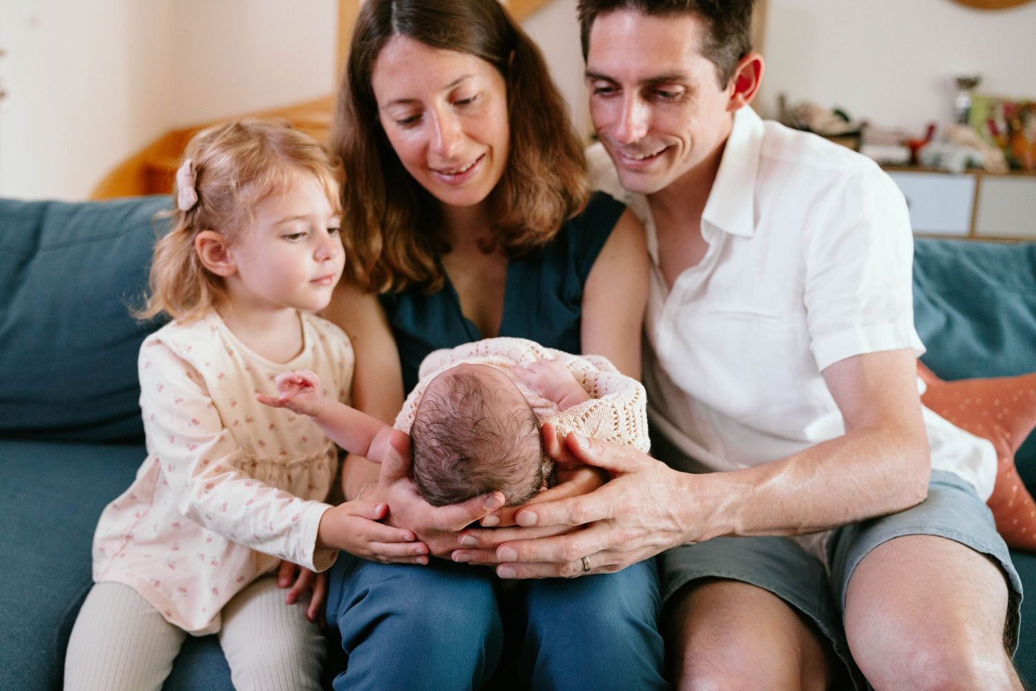 Happy parents sit on blue sofa holding newborn baby while curious toddler daughter looks closely.