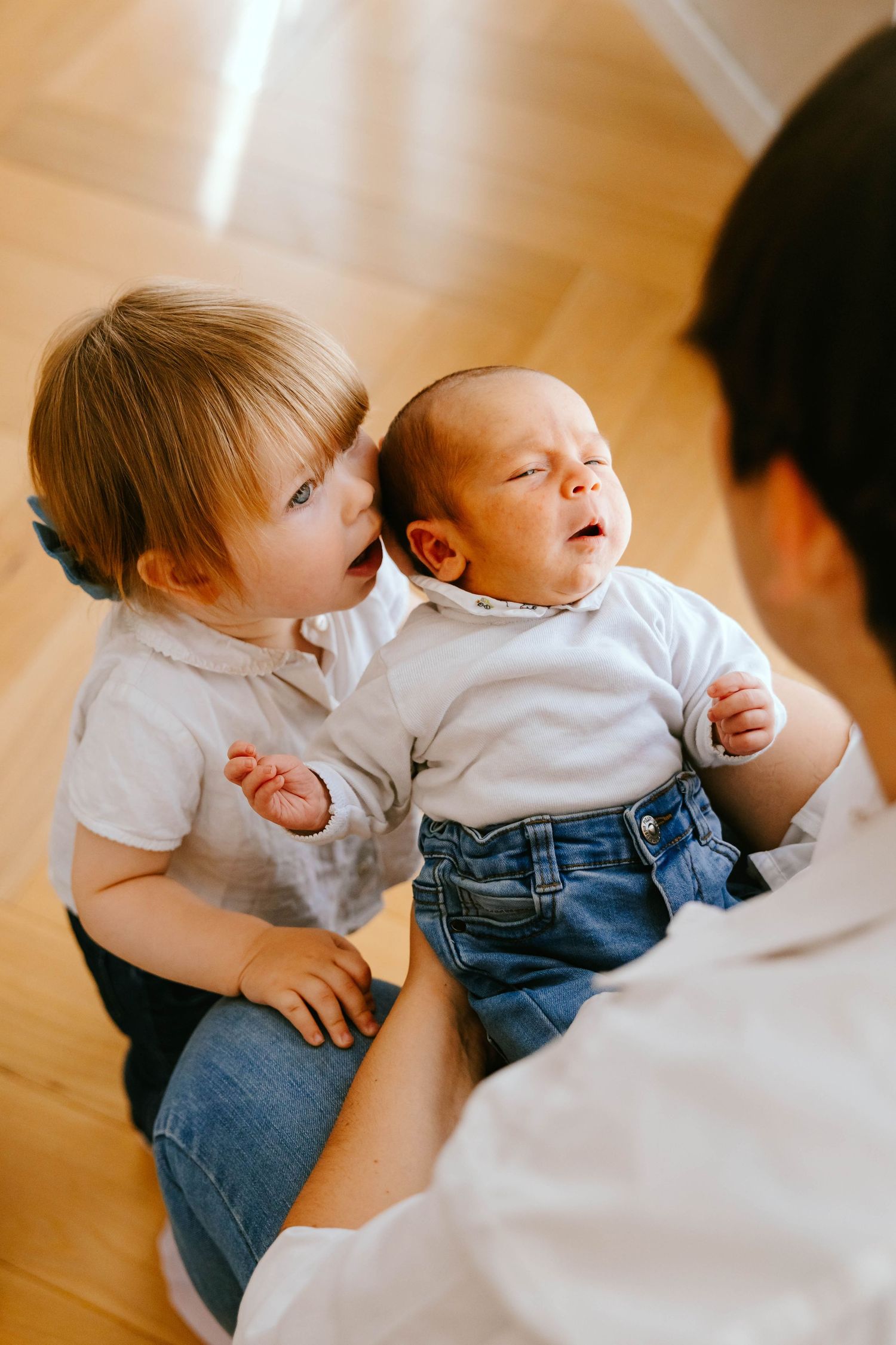 Curious toddler sibling looking at crying newborn baby held by mother on wooden floor.