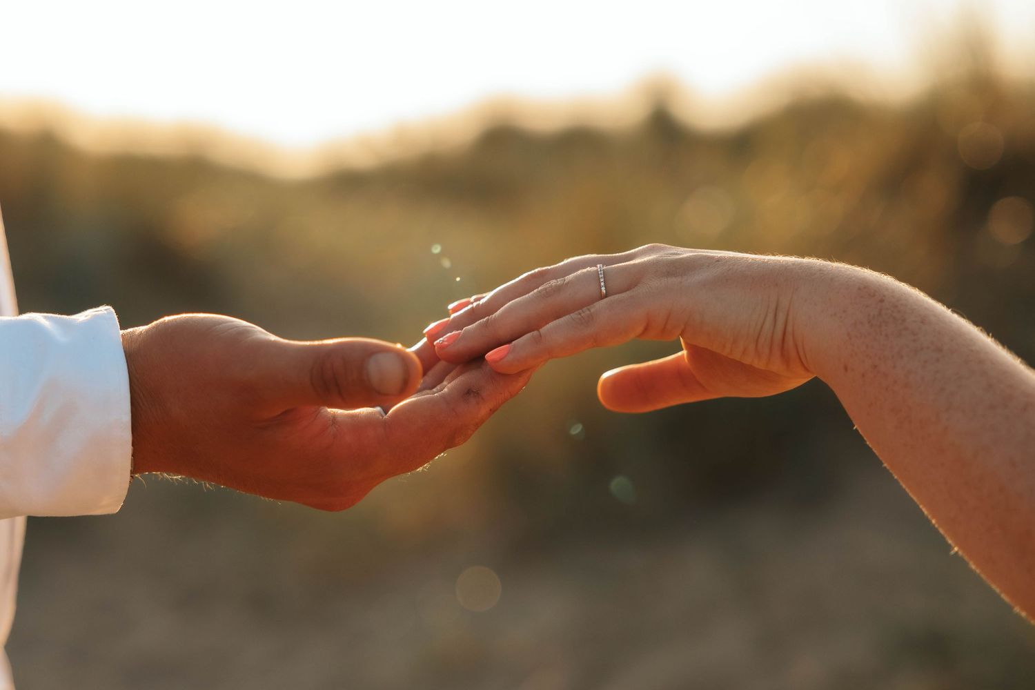 Close-up of bride and groom gently touching hands with sparkling wedding ring in warm golden sunset bokeh.