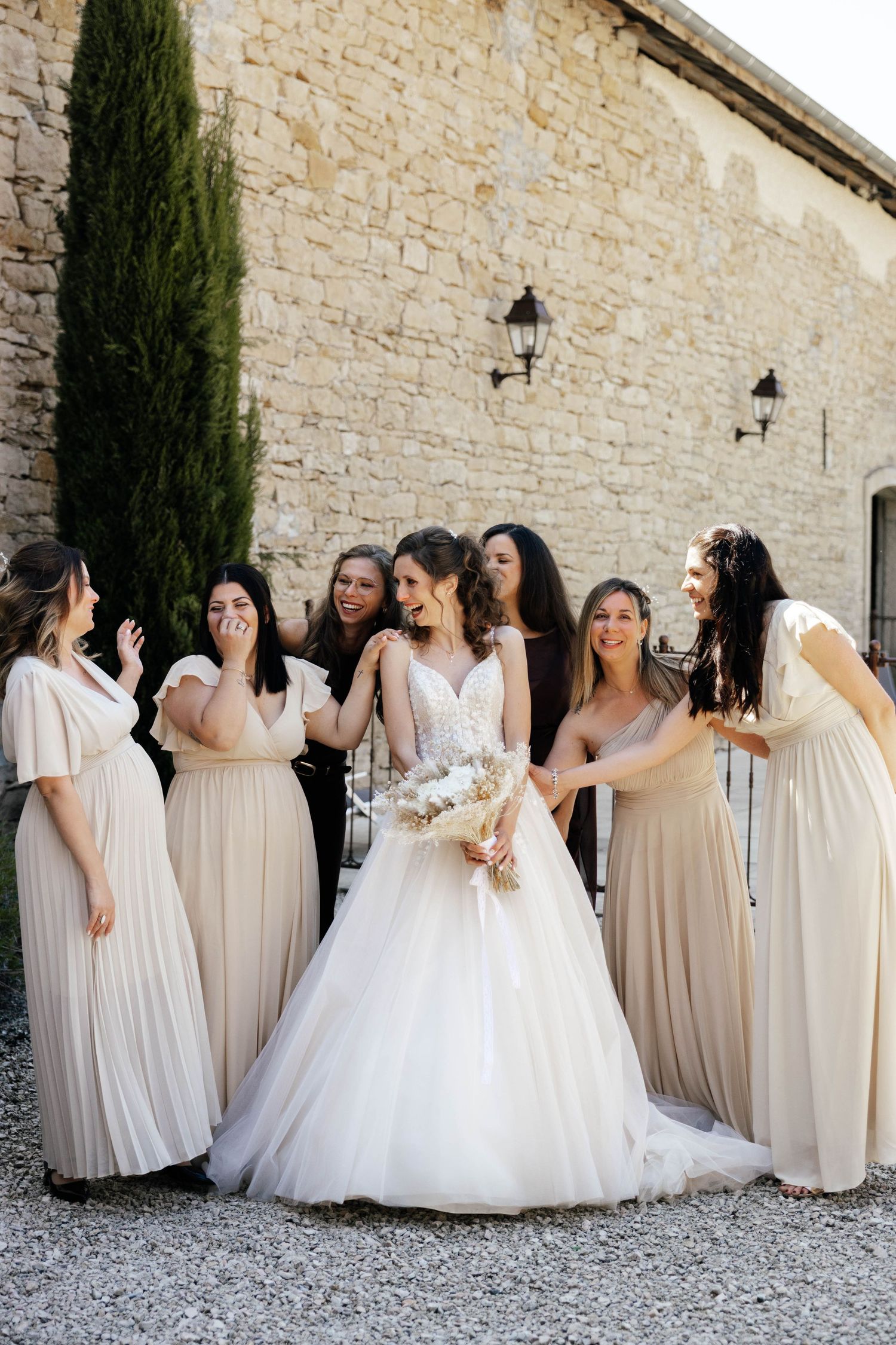 Bride in white ball gown smiles with bridesmaids in beige dresses outside a stone building.