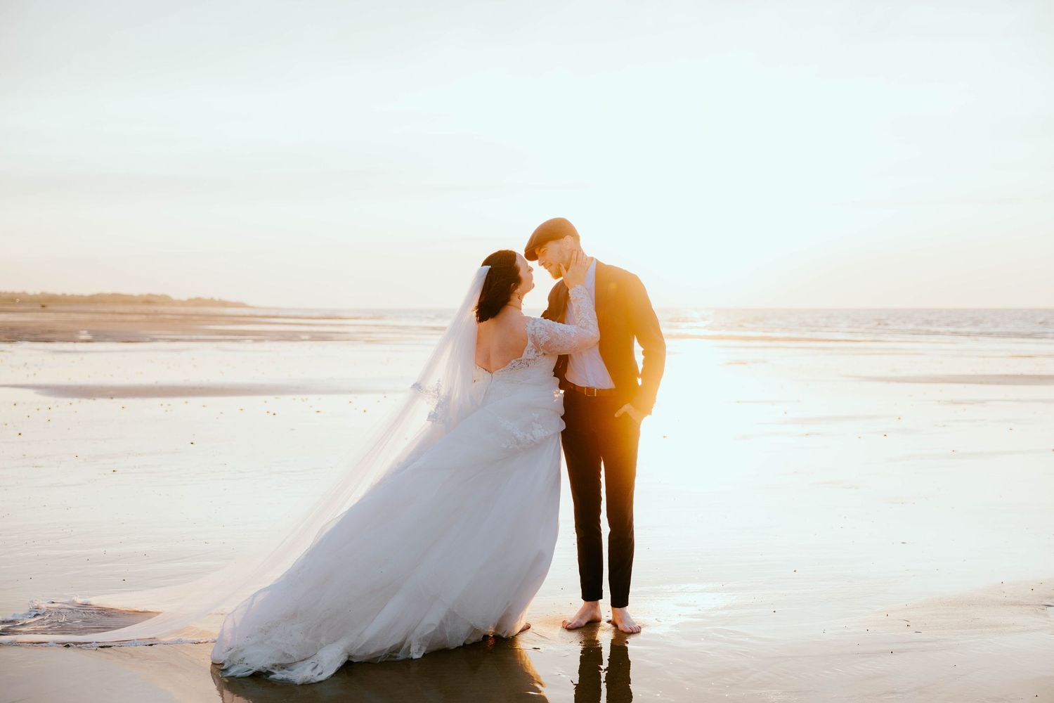 Bride and groom sharing a romantic moment on a sunlit beach, with waves reflecting golden sunset light.