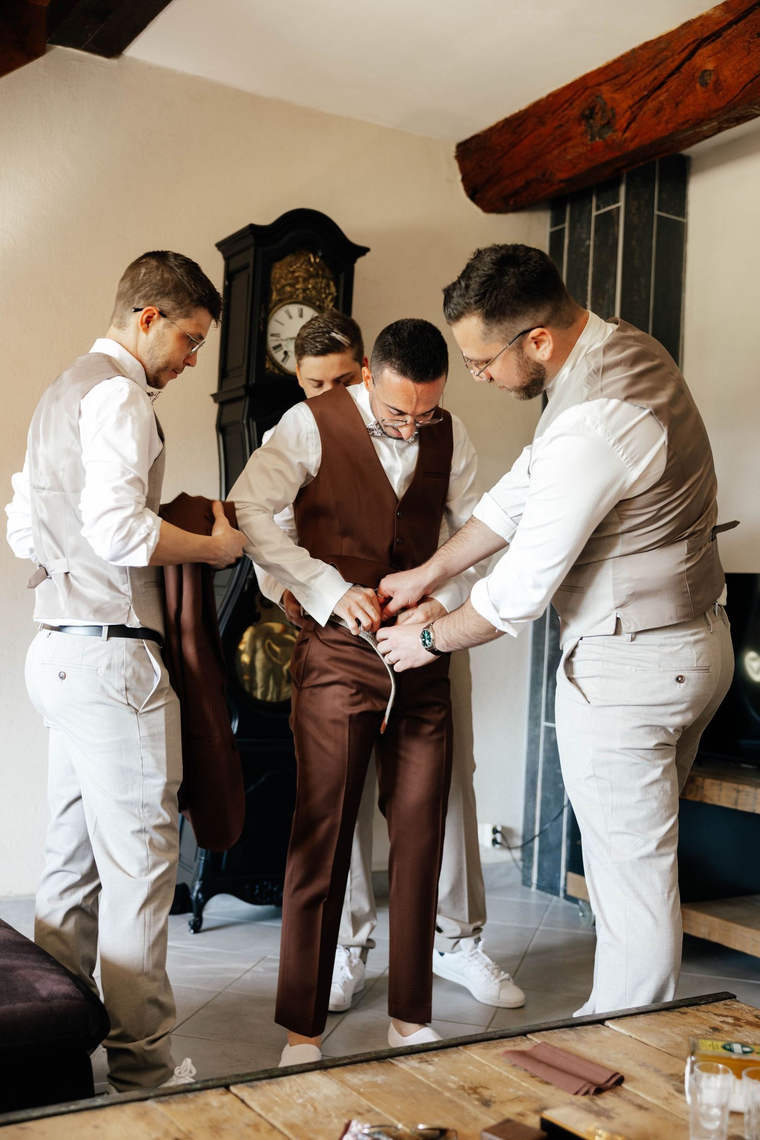 Groomsmen helping groom button his brown vest while getting ready indoors near a vintage grandfather clock.