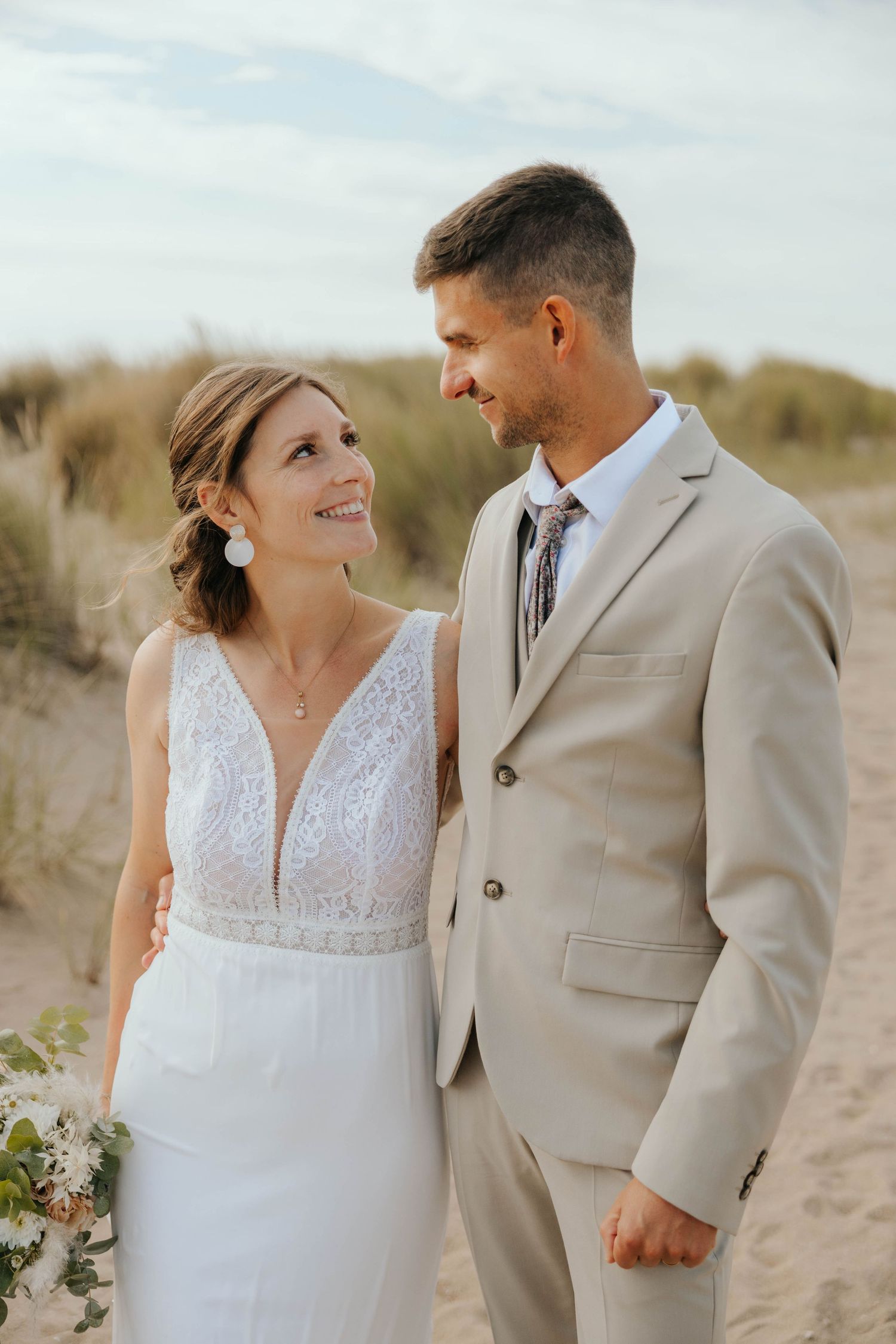 Bride and groom smiling at each other on sandy beach dunes.