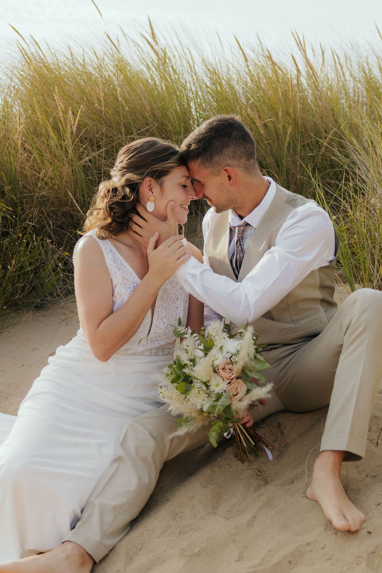 Bride and groom sitting in sand dunes touching foreheads romantically.