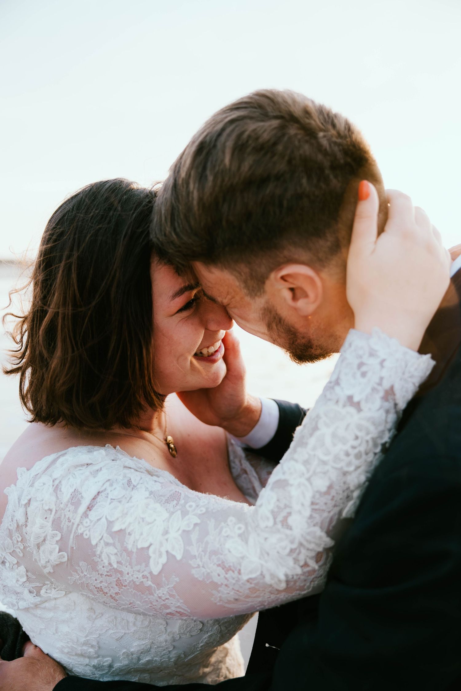 Happy bride in lace dress laughing as groom pulls her close in a tender outdoor embrace.