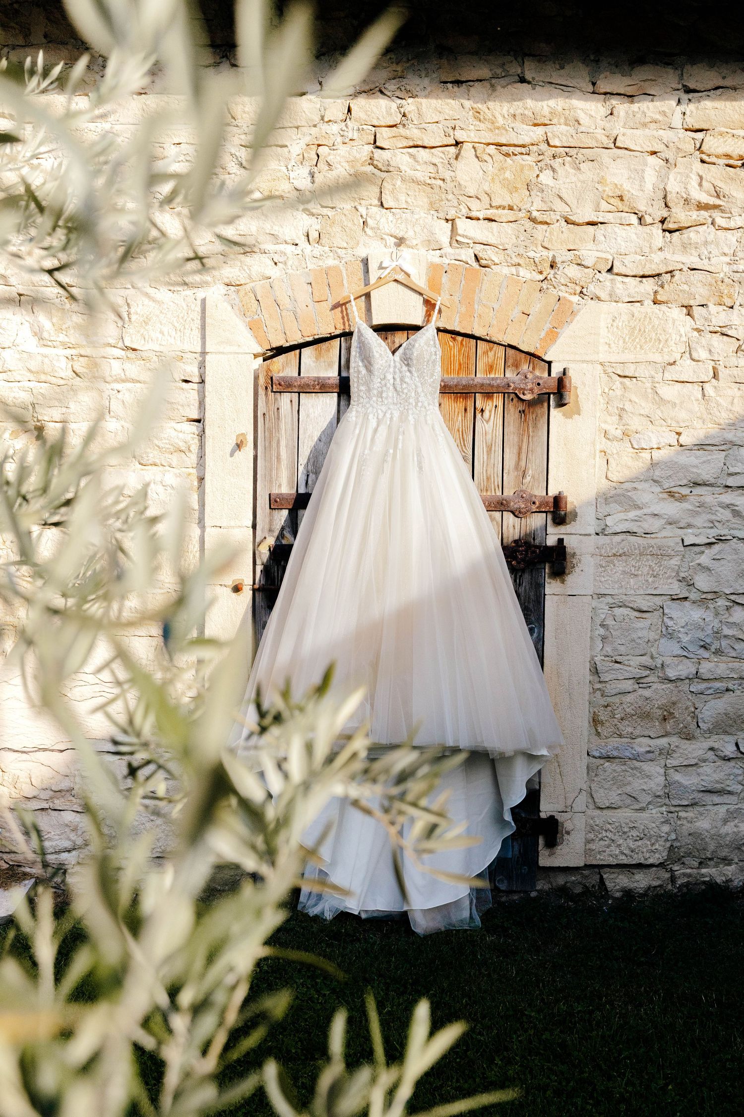 Elegant white wedding dress hanging on wooden shutters against a rustic stone wall with olive tree branches in foreground.