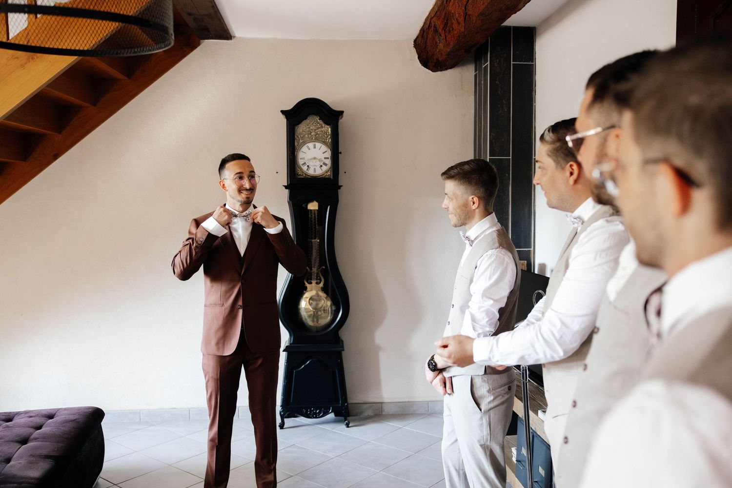 Groom in burgundy suit adjusting bow tie with groomsmen in white suits near vintage grandfather clock.