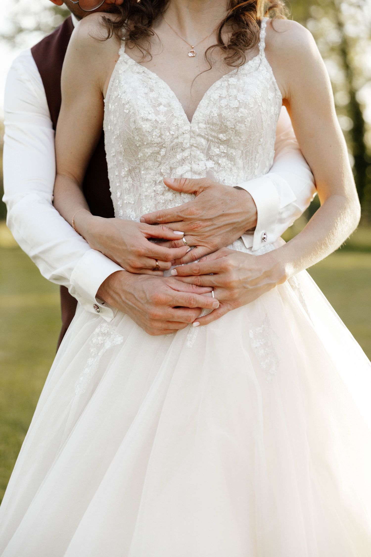 Groom embracing bride from behind, both wearing wedding rings, bride in lace gown.