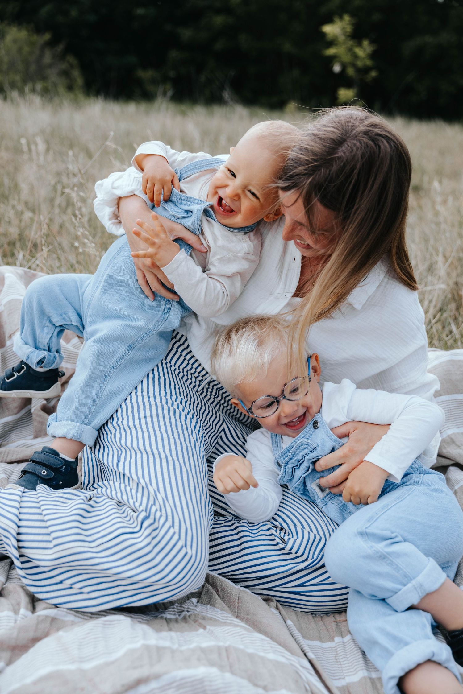 A mother sits on a blanket outdoors, joyfully cuddling her two young toddlers in blue outfits amid a grassy field.