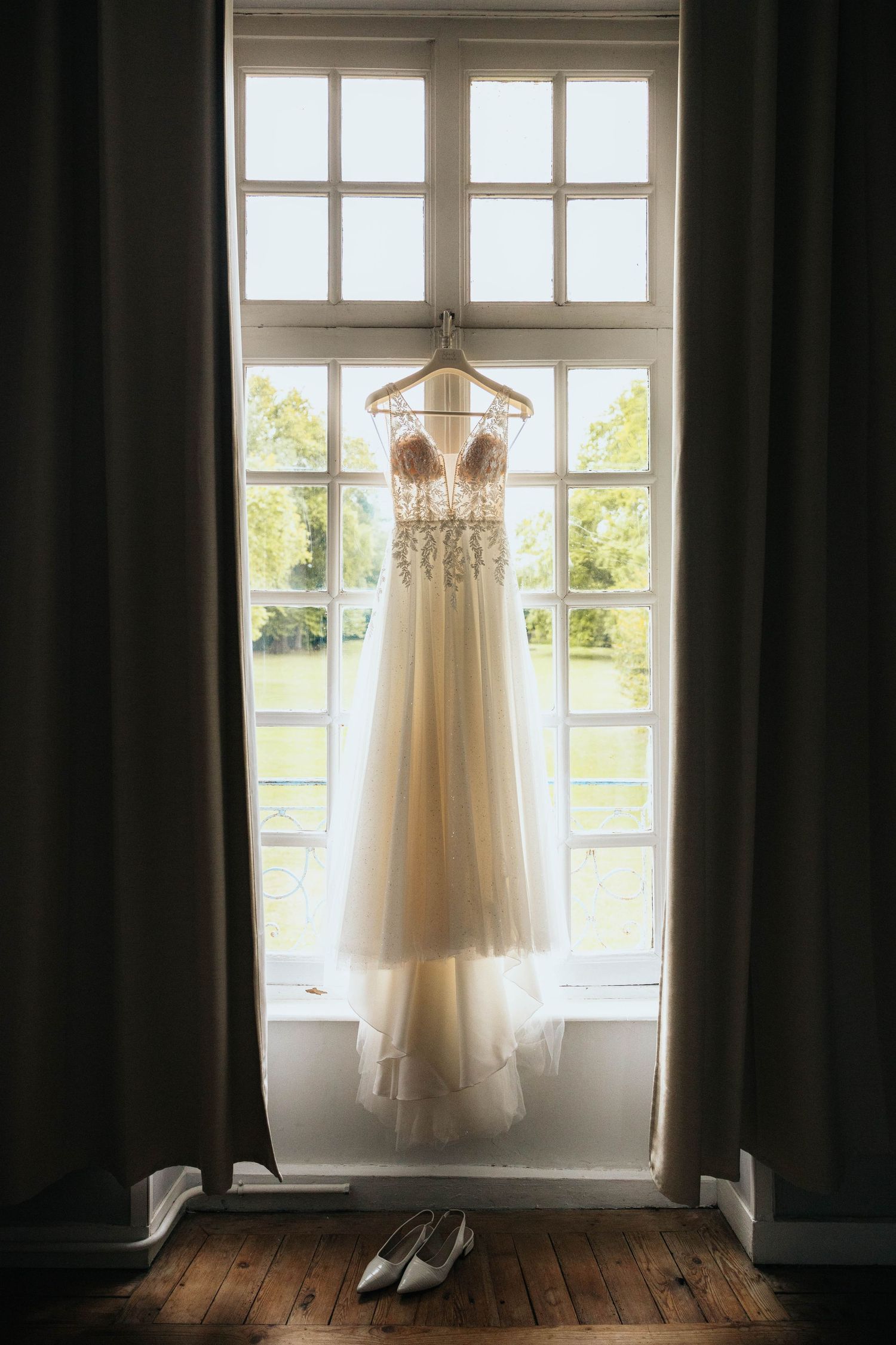 Elegant beaded wedding dress hanging in a sunlit window with white bridal shoes below on hardwood floor.