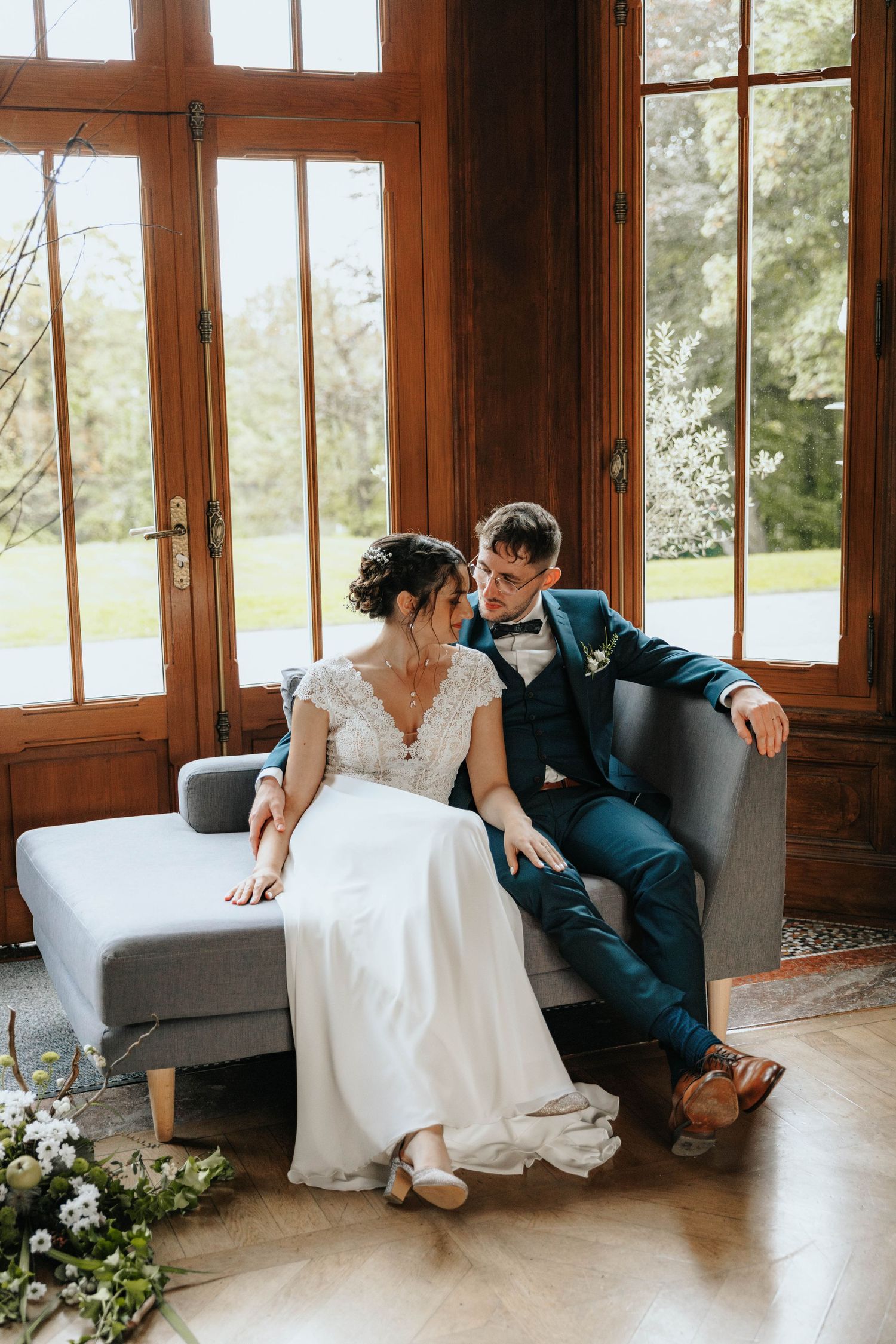 Bride and groom sitting together on a gray sofa in a wood-paneled room with large windows on their wedding day.