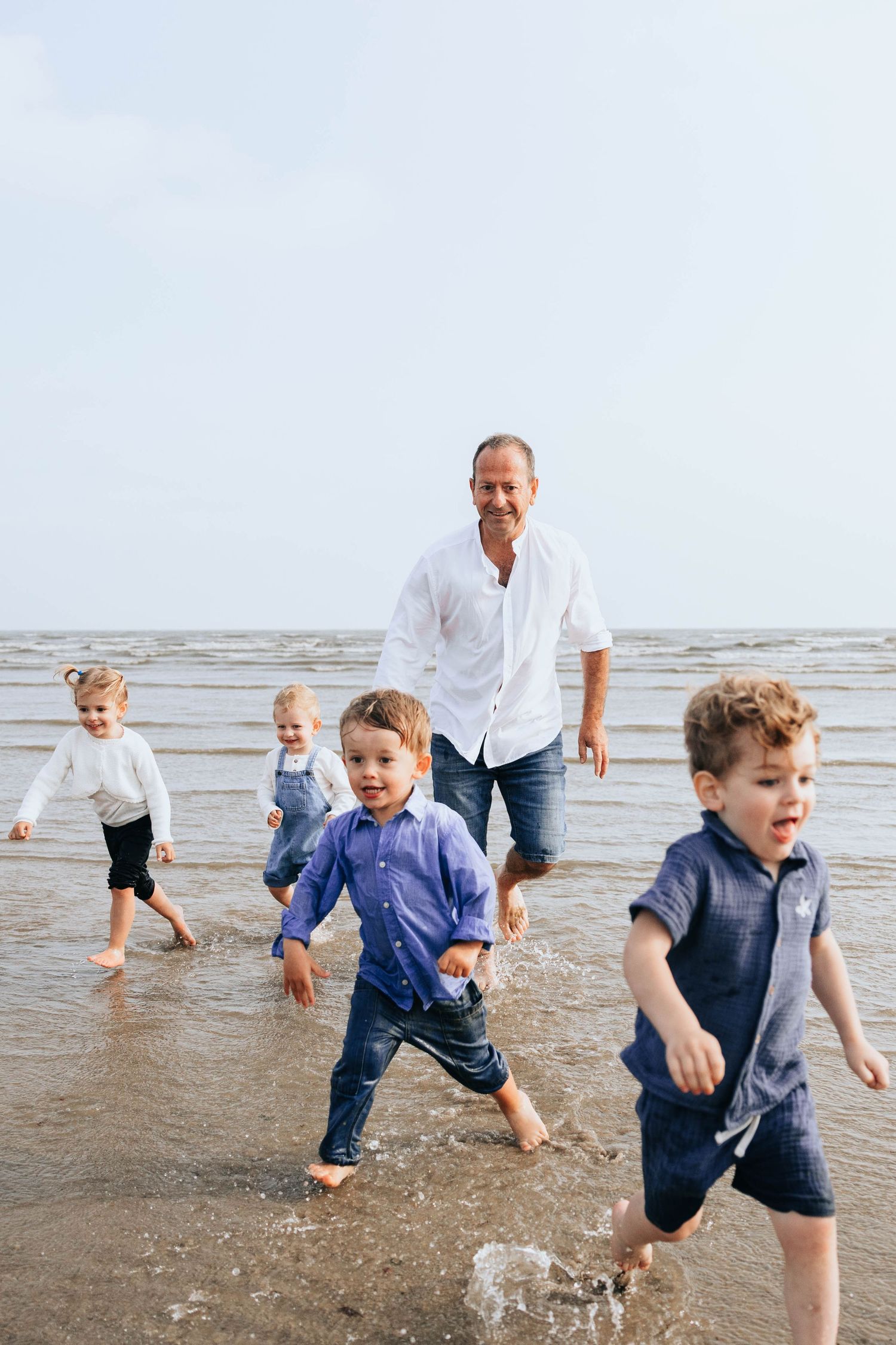 Father in white shirt plays with four young children splashing in shallow ocean waves.