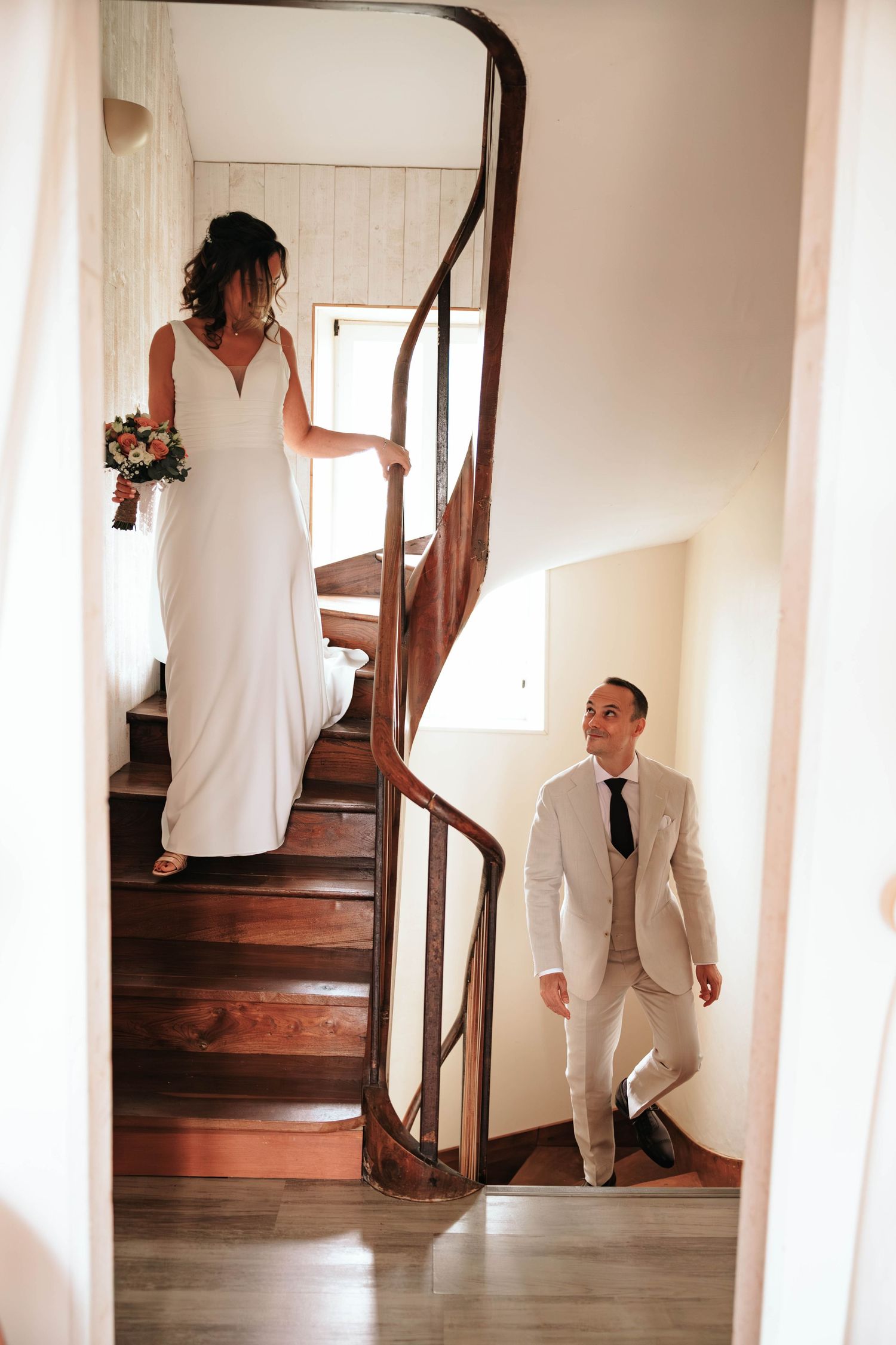 Groom in beige suit smiling up at bride descending wooden spiral staircase holding bridal bouquet.