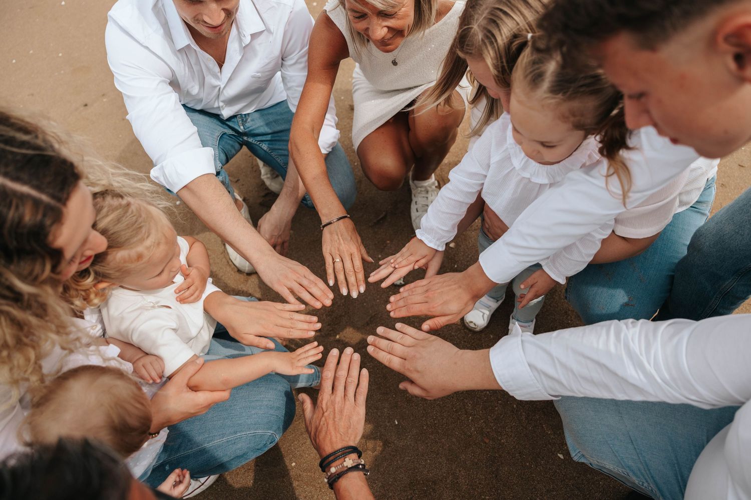 Group of adults and children forming a circle with hands together over sandy ground.