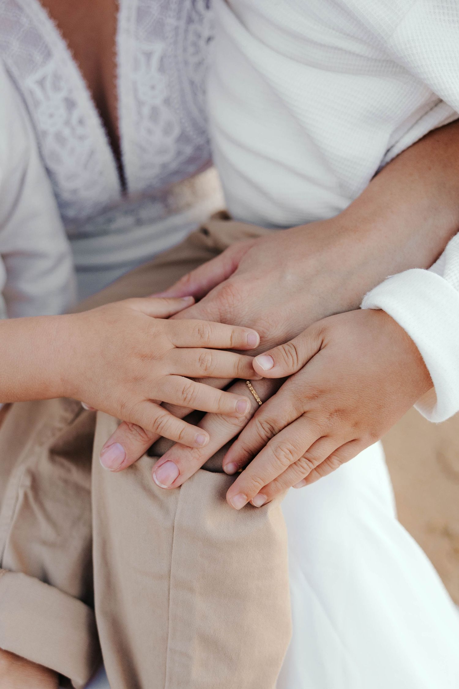 Close-up of two people holding hands tenderly, one wearing a ring, dressed in white and beige clothing.