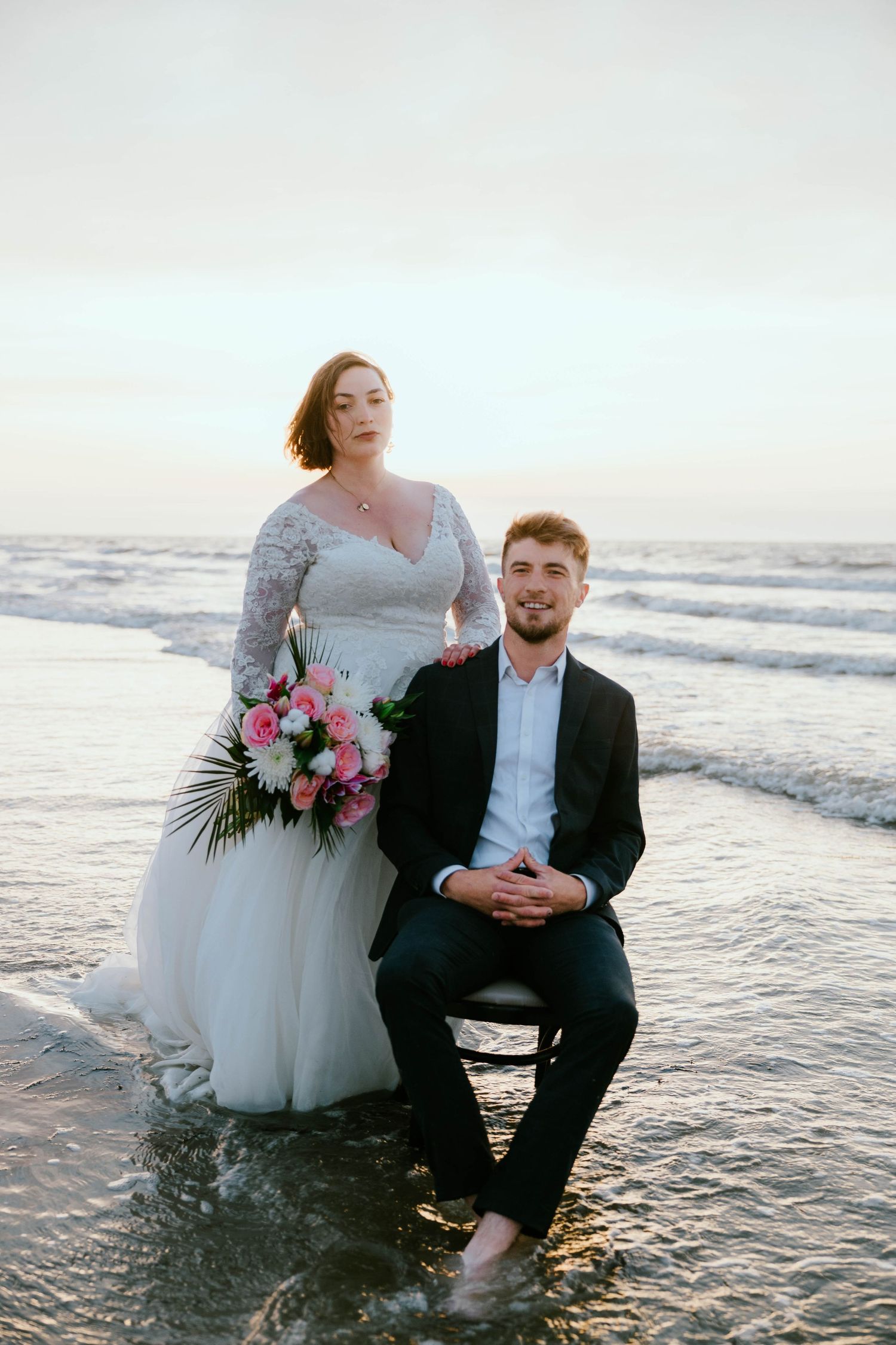 Bride in lace gown holding pink bouquet stands beside groom in black suit sitting on beach at sunset.