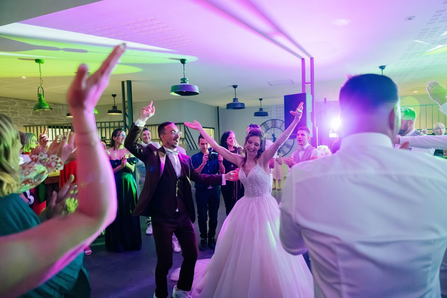 Bride and groom dance joyfully with guests under colorful purple and pink lights at lively wedding reception.