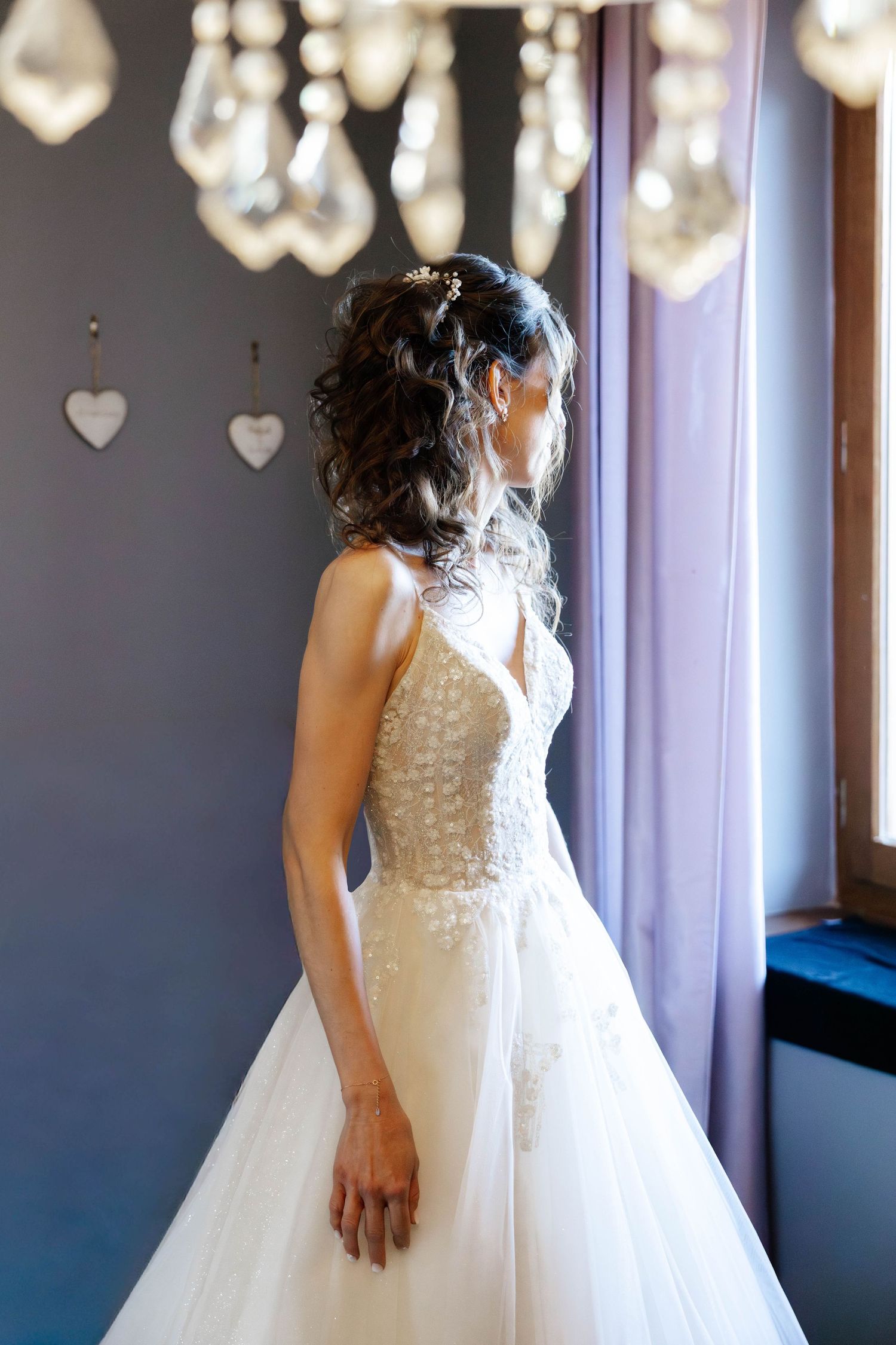 Bride in elegant lace wedding gown standing by window with crystal chandelier overhead.