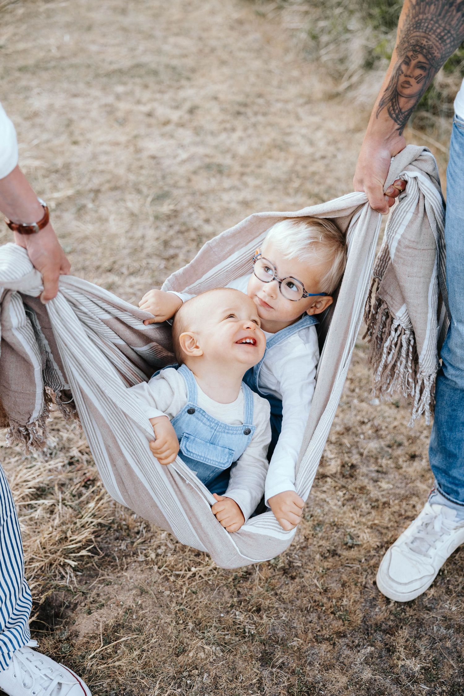 Two young children sitting together in a hammock outdoors, smiling joyfully while adults hold the sides.