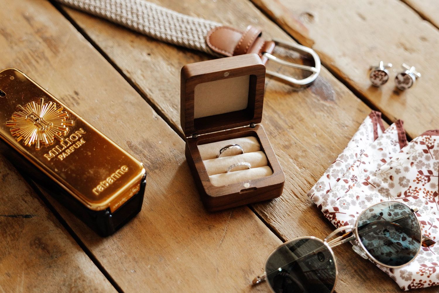 Wedding rings in a wooden box beside sunglasses, a gold cologne box, and a floral pocket square on a wooden surface.