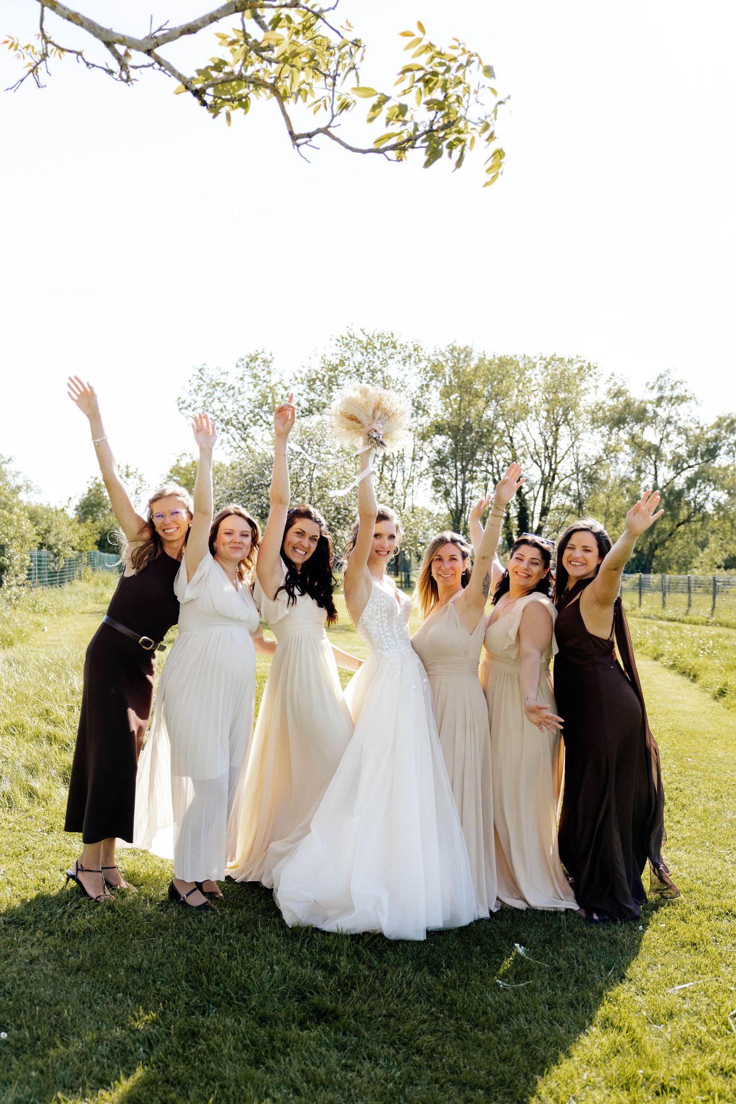Bride with bridesmaids in neutral gowns celebrating outdoors with arms raised joyfully.