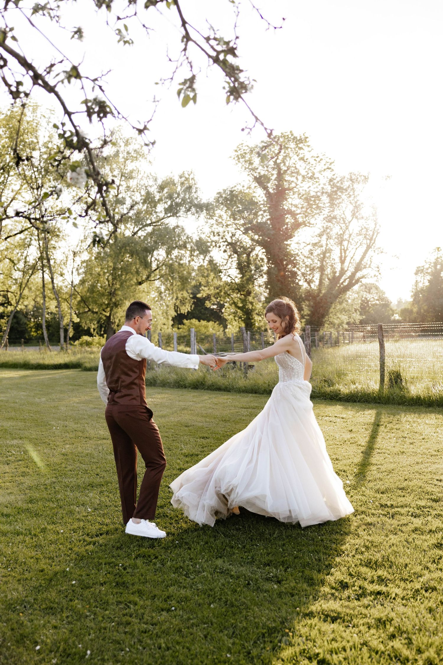 Bride and groom dancing together on a sunlit green lawn surrounded by trees.