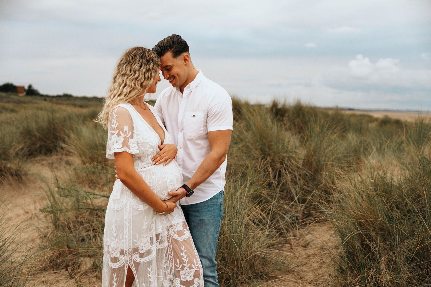 Expecting couple embracing tenderly in sand dunes, woman in white lace maternity dress with hands on belly.