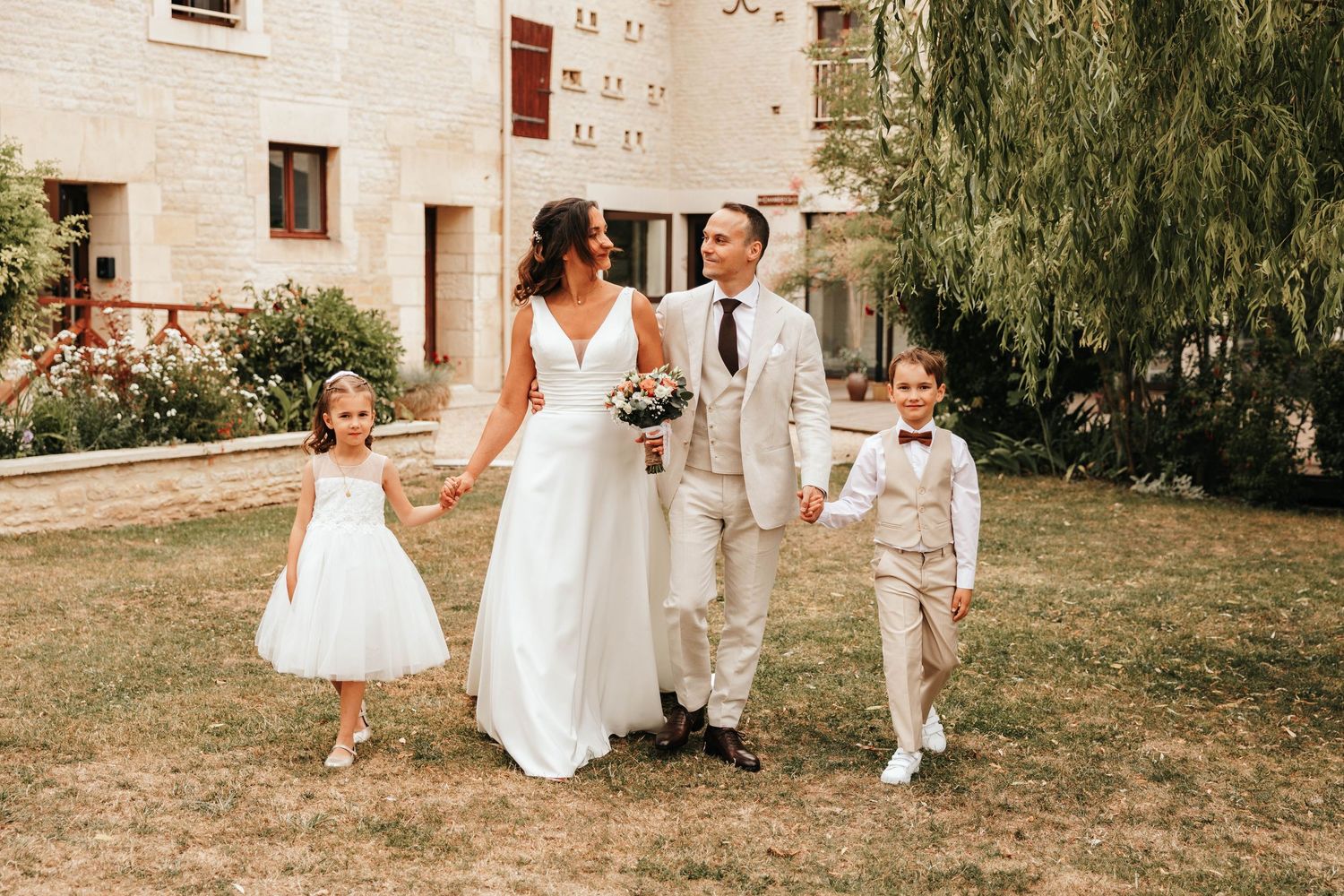 Happy wedding couple walking outdoors with flower girl and ring bearer on sunny day at chateau venue.