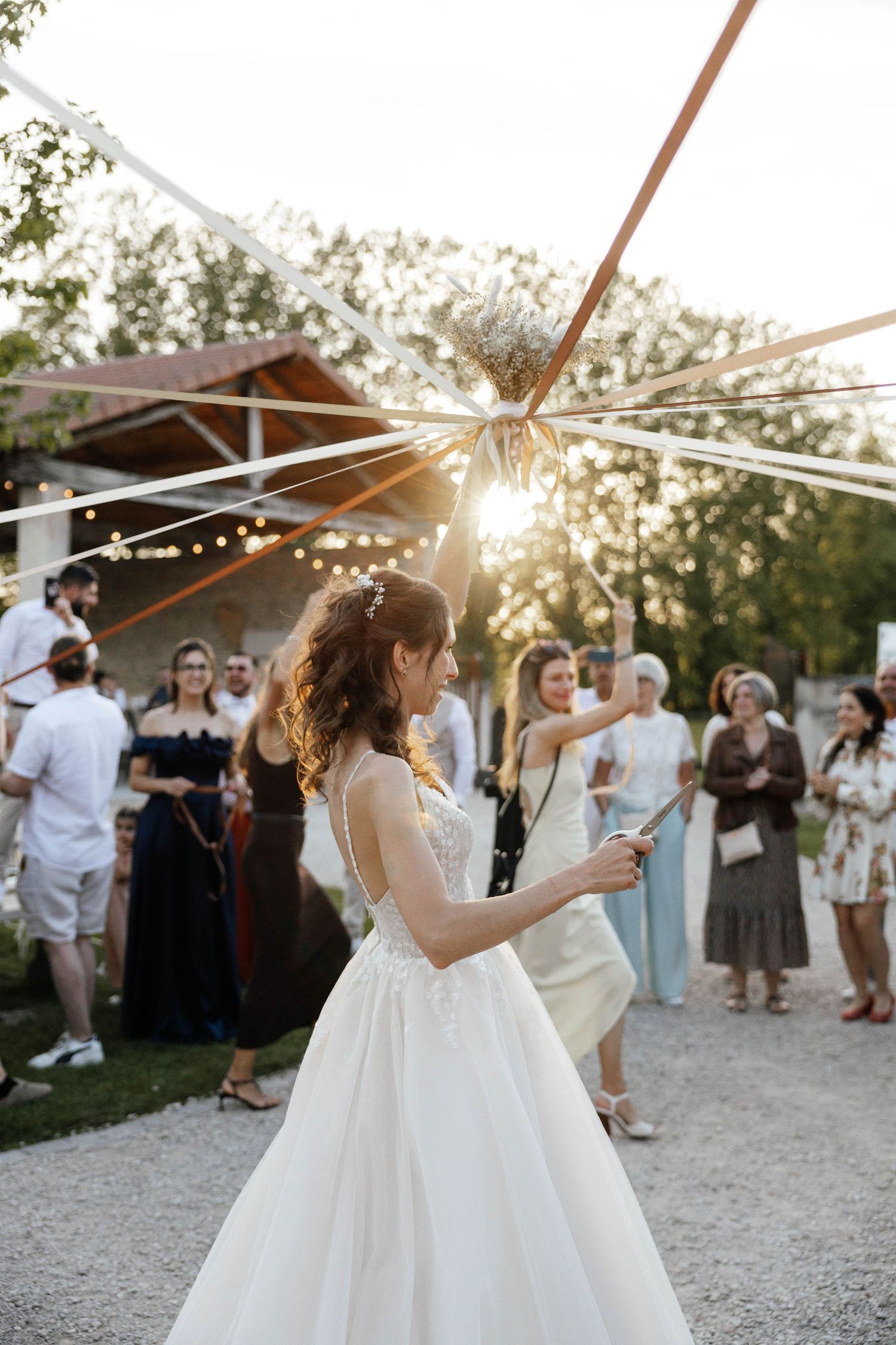 Bride in white lace gown holding sparkler at outdoor sunset wedding reception.