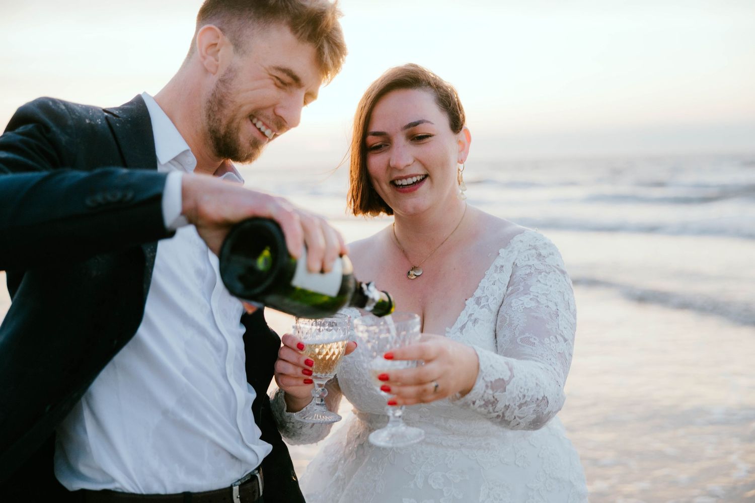Bride and groom celebrating on beach, groom pouring champagne into glasses at sunset wedding.