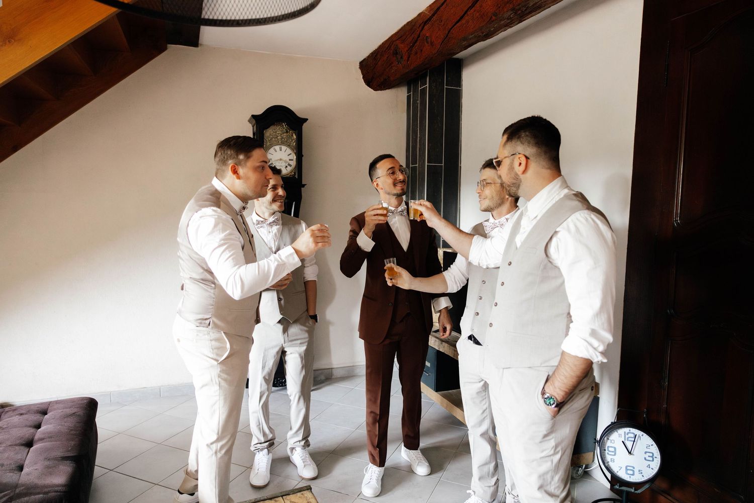 Groom in brown suit toasts with groomsmen in white suits indoors near a grandfather clock at a wedding.