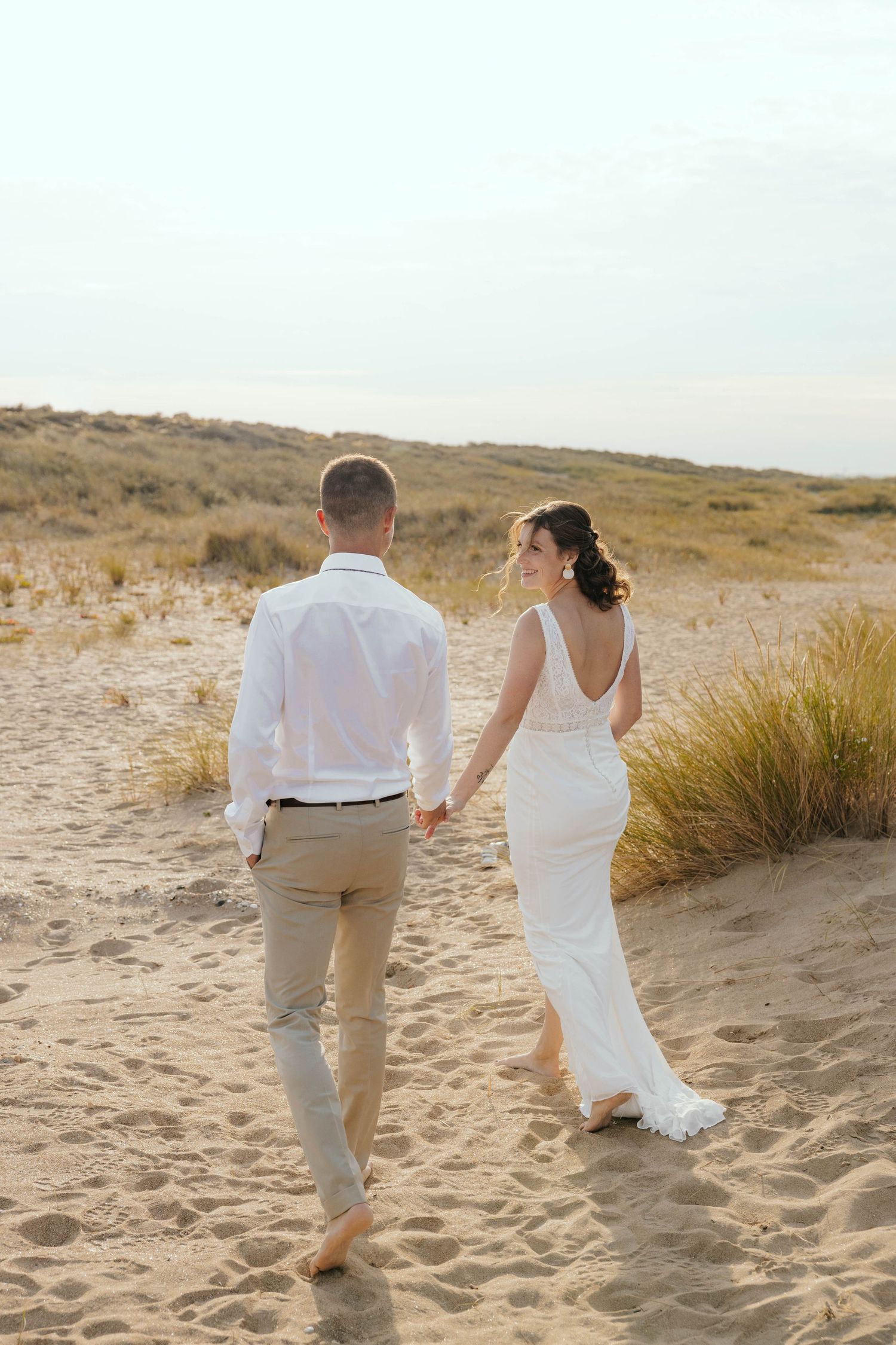 Bride and groom walking hand in hand barefoot on beach sand.