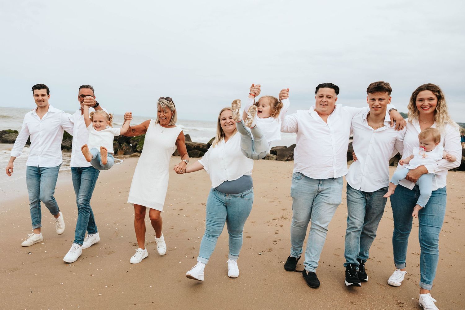 Joyful family in matching white and jeans walking along sandy beach with arms raised.