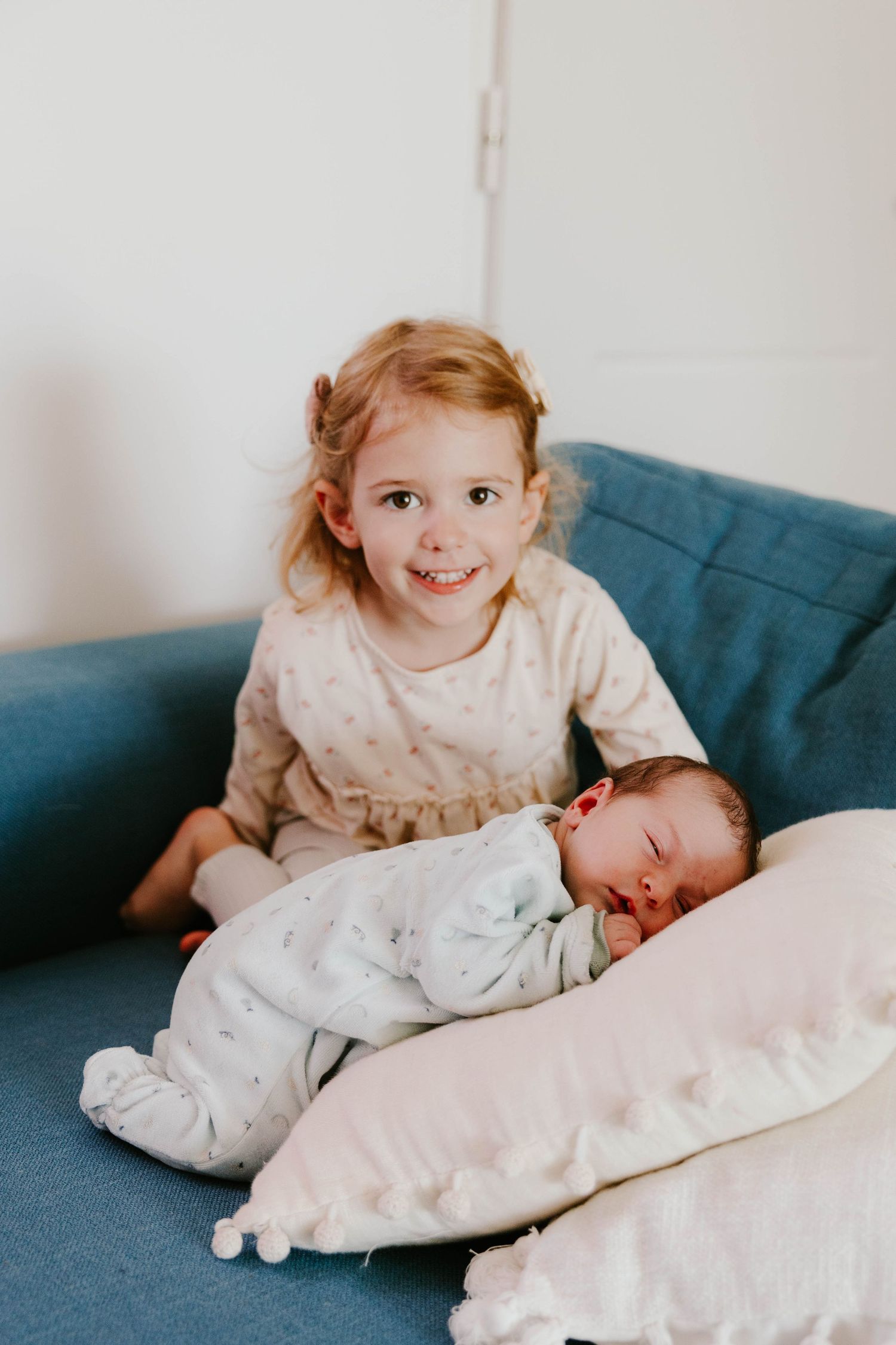 Smiling toddler girl sits on blue sofa beside sleeping newborn baby sibling propped on a pillow.