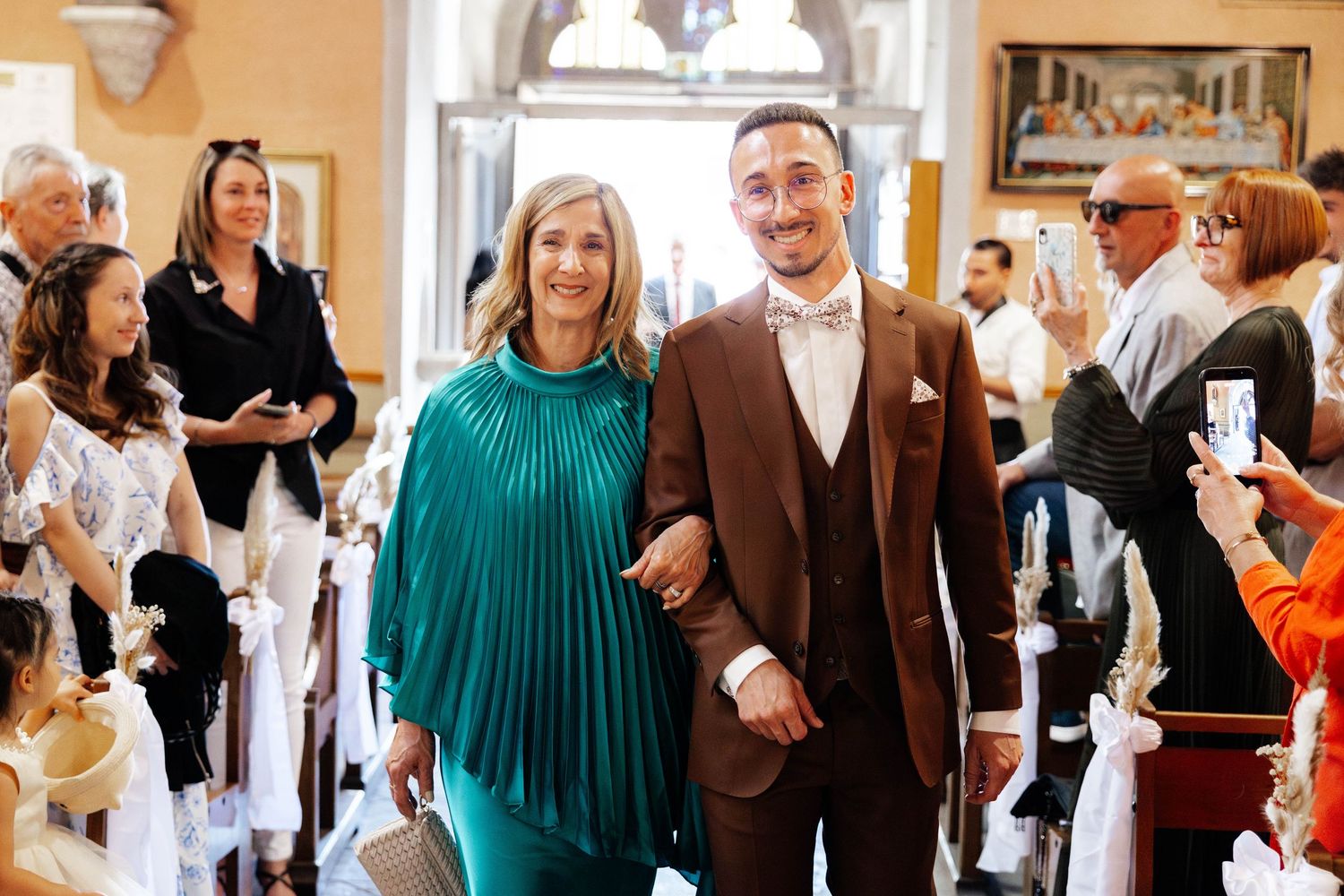 A groom in a brown suit and a woman in a teal dress walk hand in hand down a church aisle at a wedding.