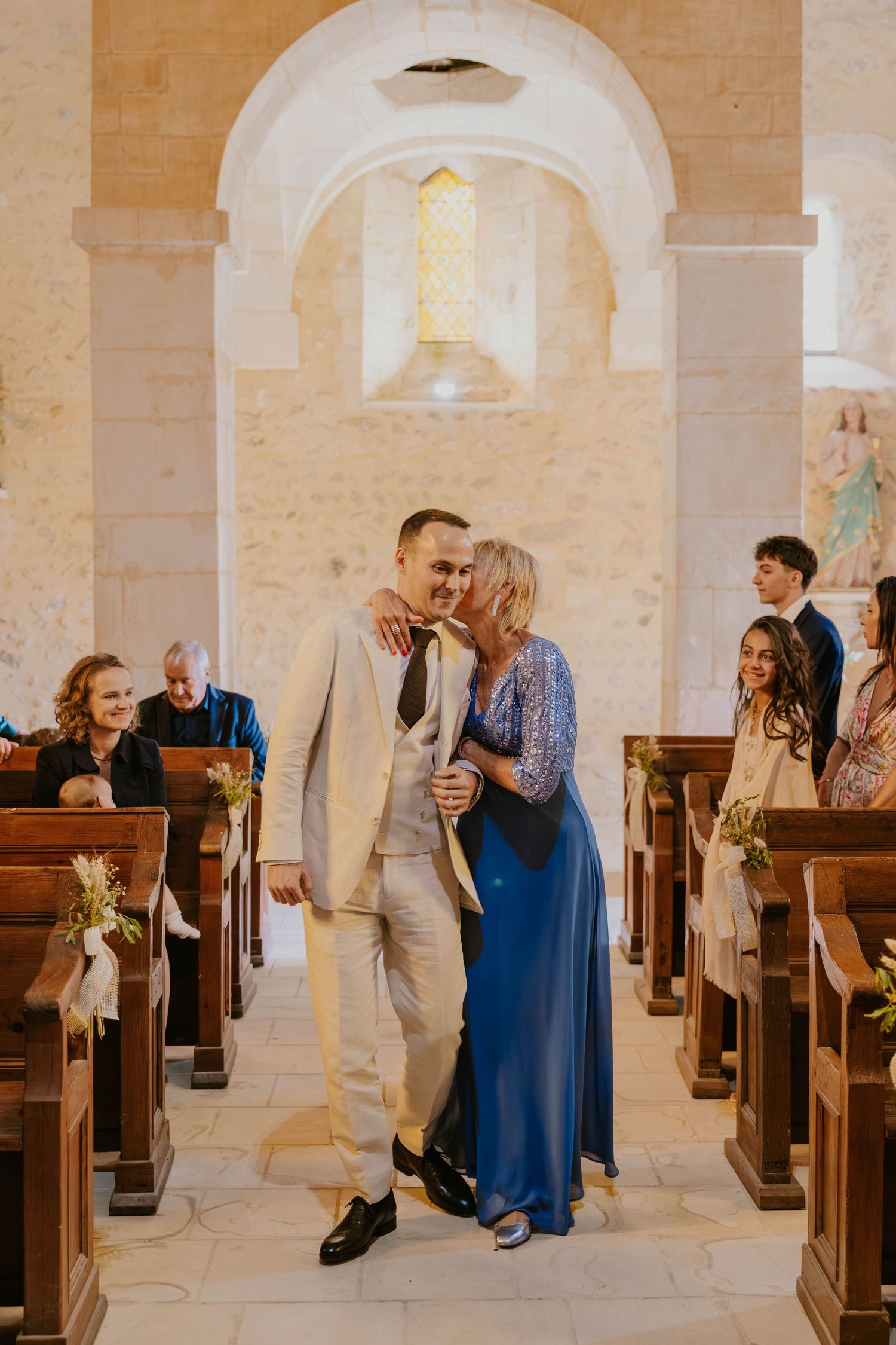 Groom walks down church aisle with a woman in a blue dress who kisses his cheek during wedding ceremony.