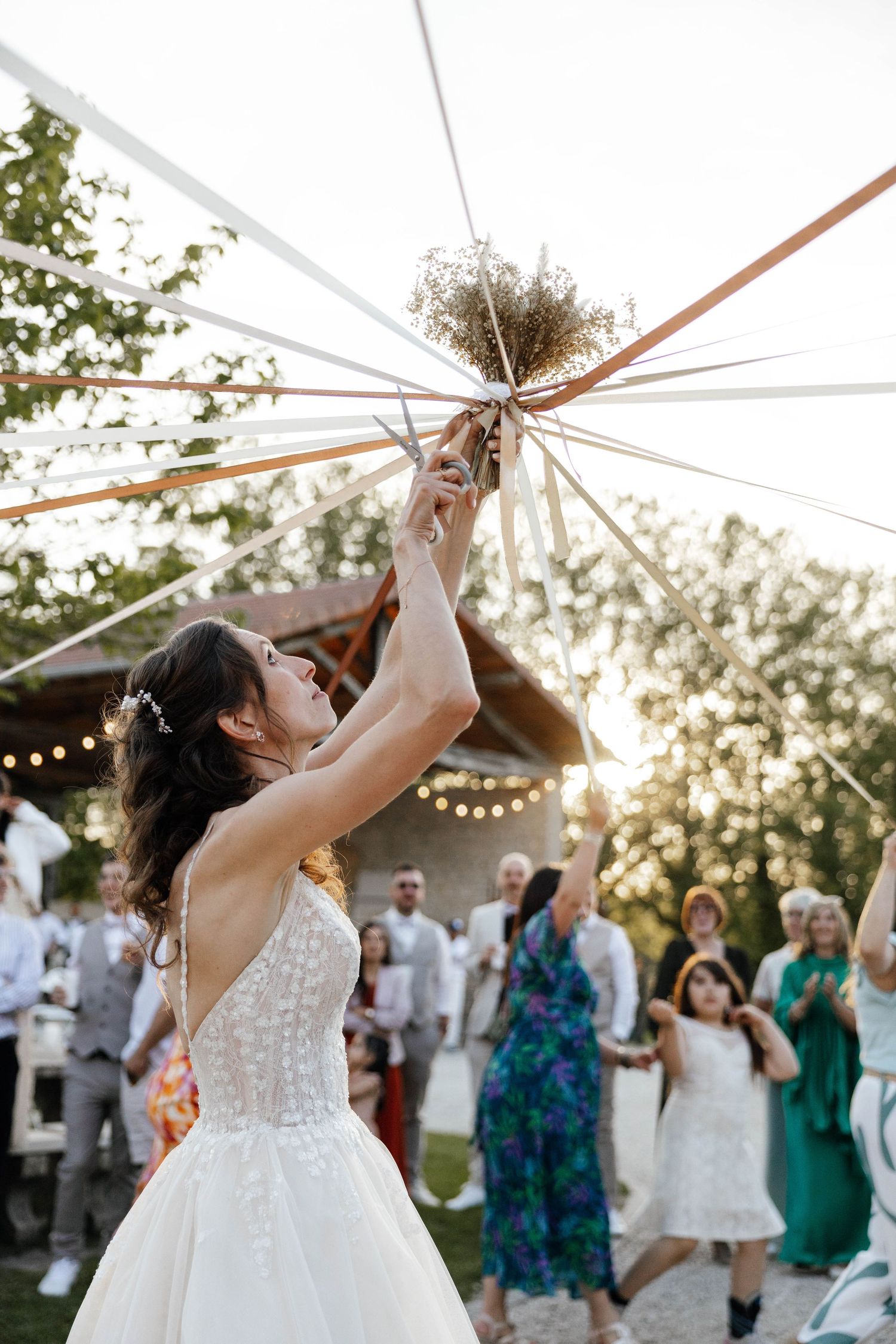 Bride in white gown tosses bouquet with ribbons at outdoor wedding reception at sunset.