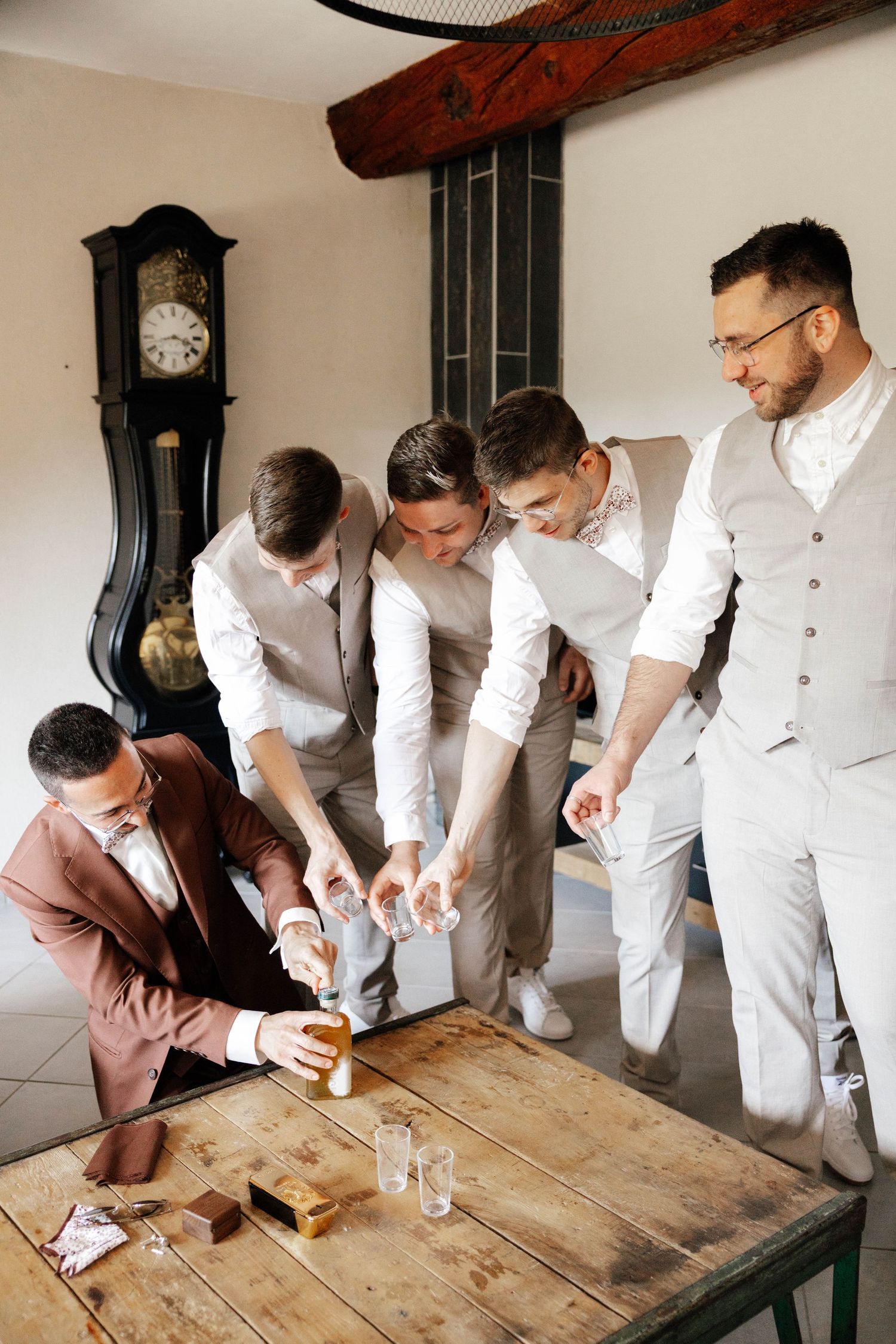 Groomsmen in elegant suits gather around a rustic wooden table, pouring whiskey shots before a wedding ceremony.