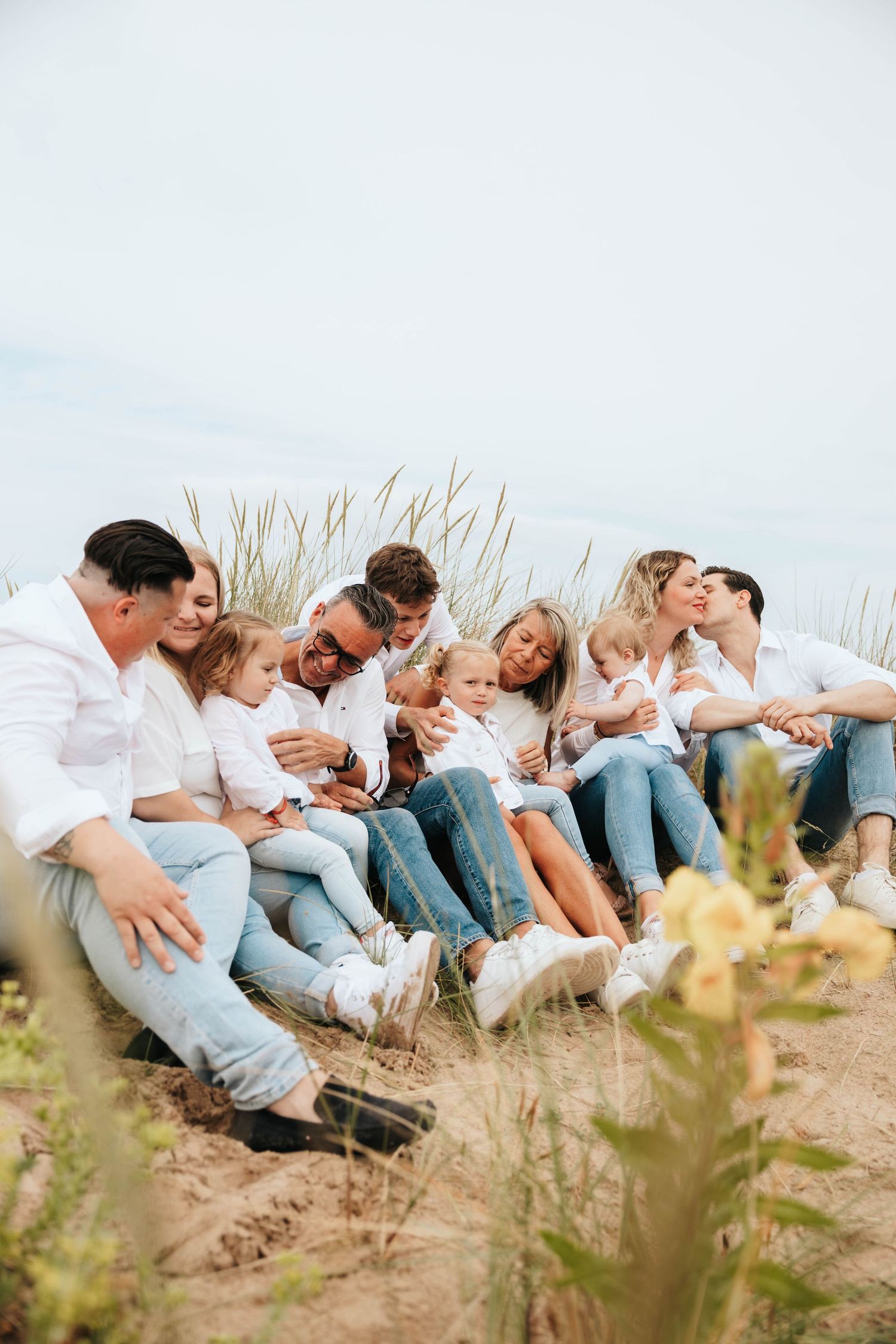 Extended family sitting together in sand dunes, cuddling young children in casual white outfits.