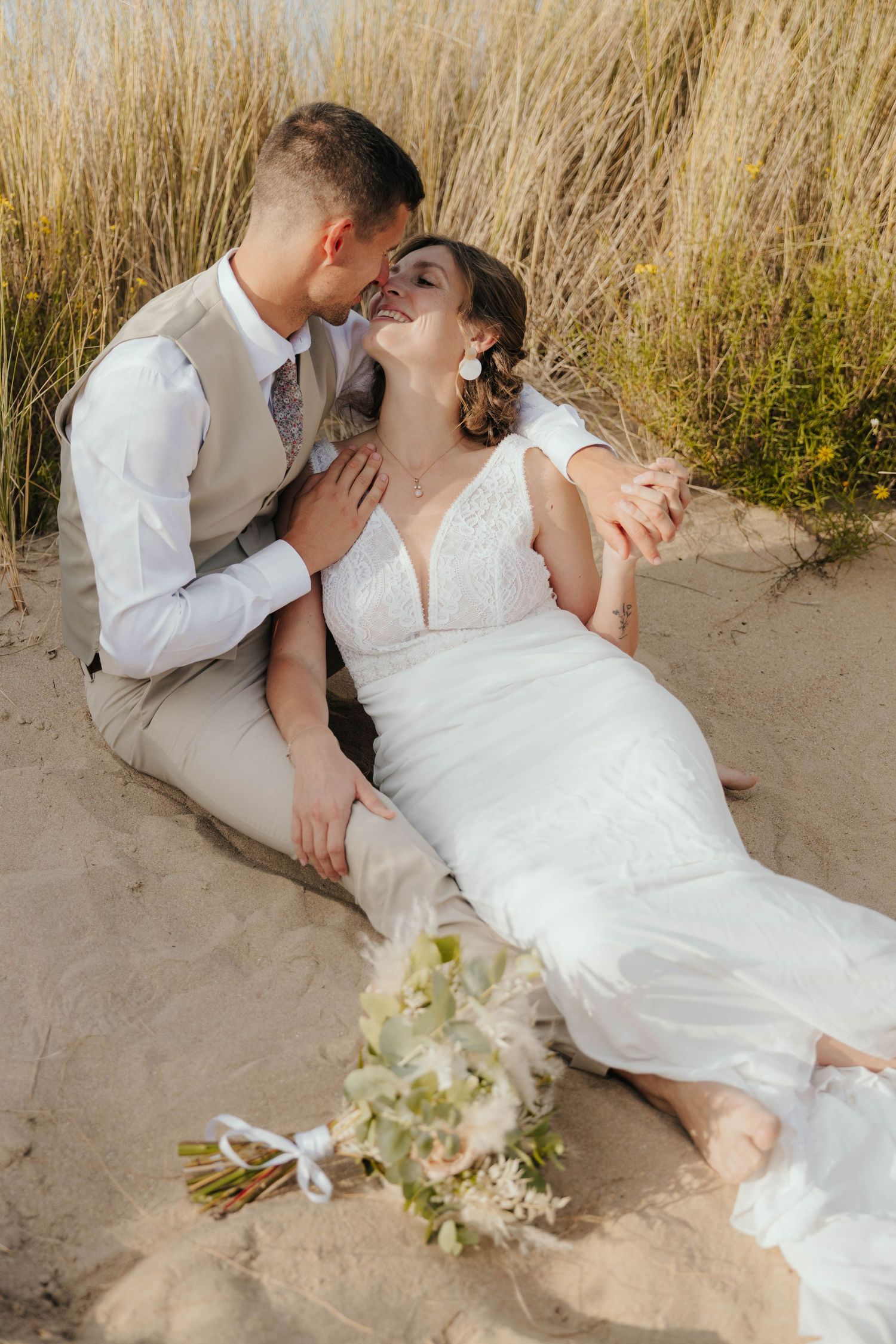 Groom kissing bride as she laughs on sandy beach dunes.