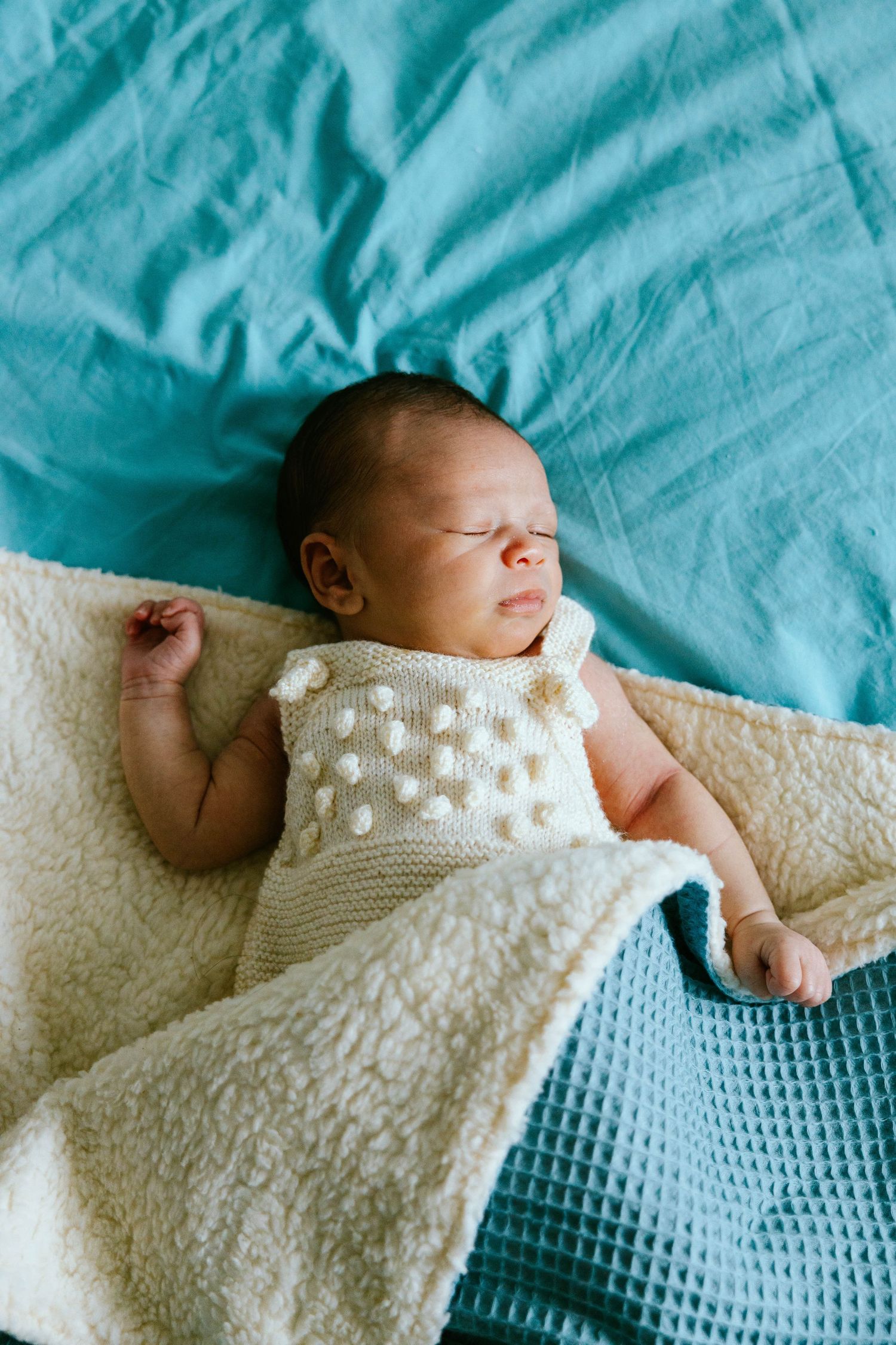 Peaceful newborn baby sleeping on teal blanket wrapped in cream knit outfit.