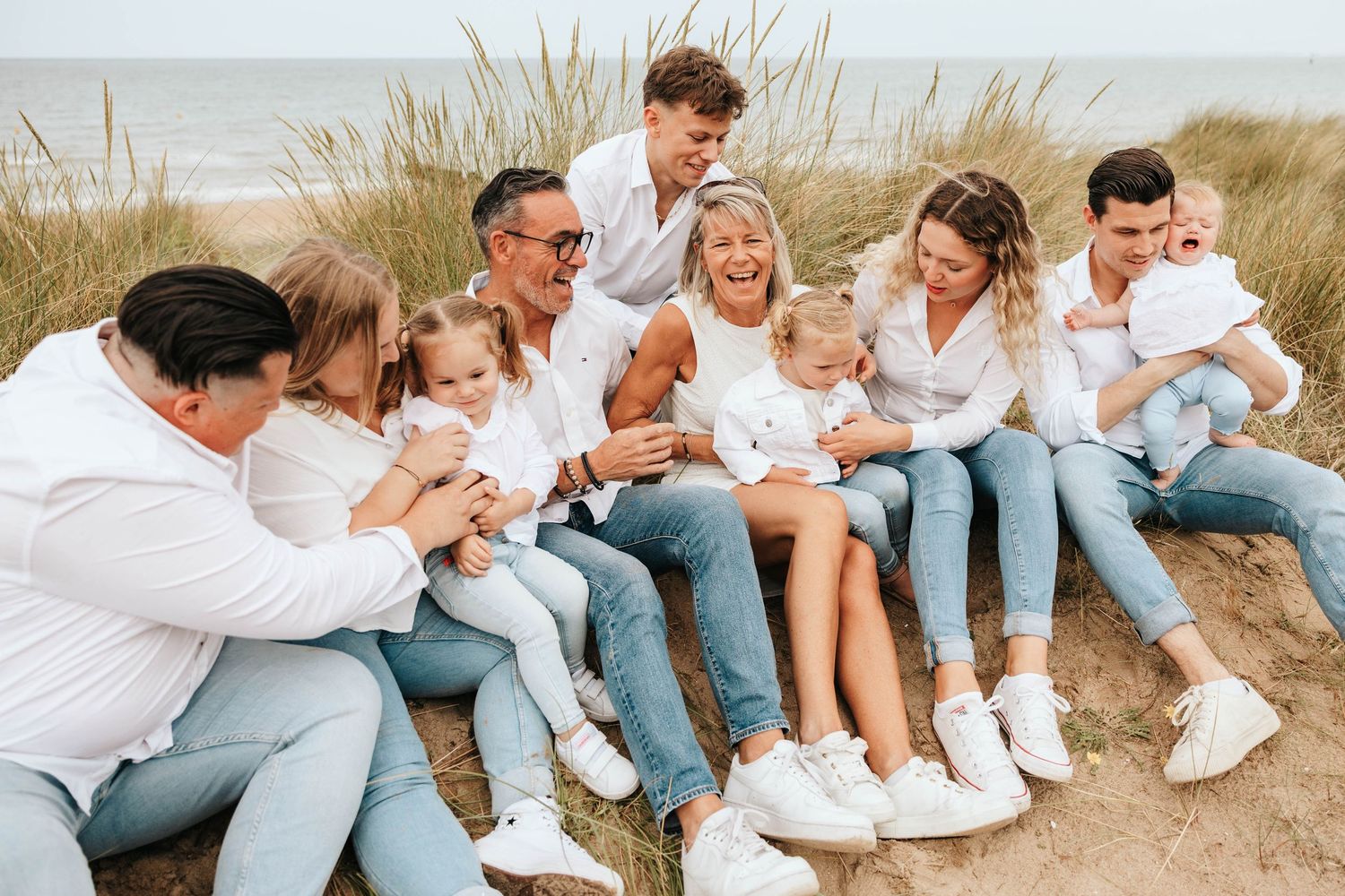 A large multigenerational family in white and denim laughs together while seated on a sunny sand dune.