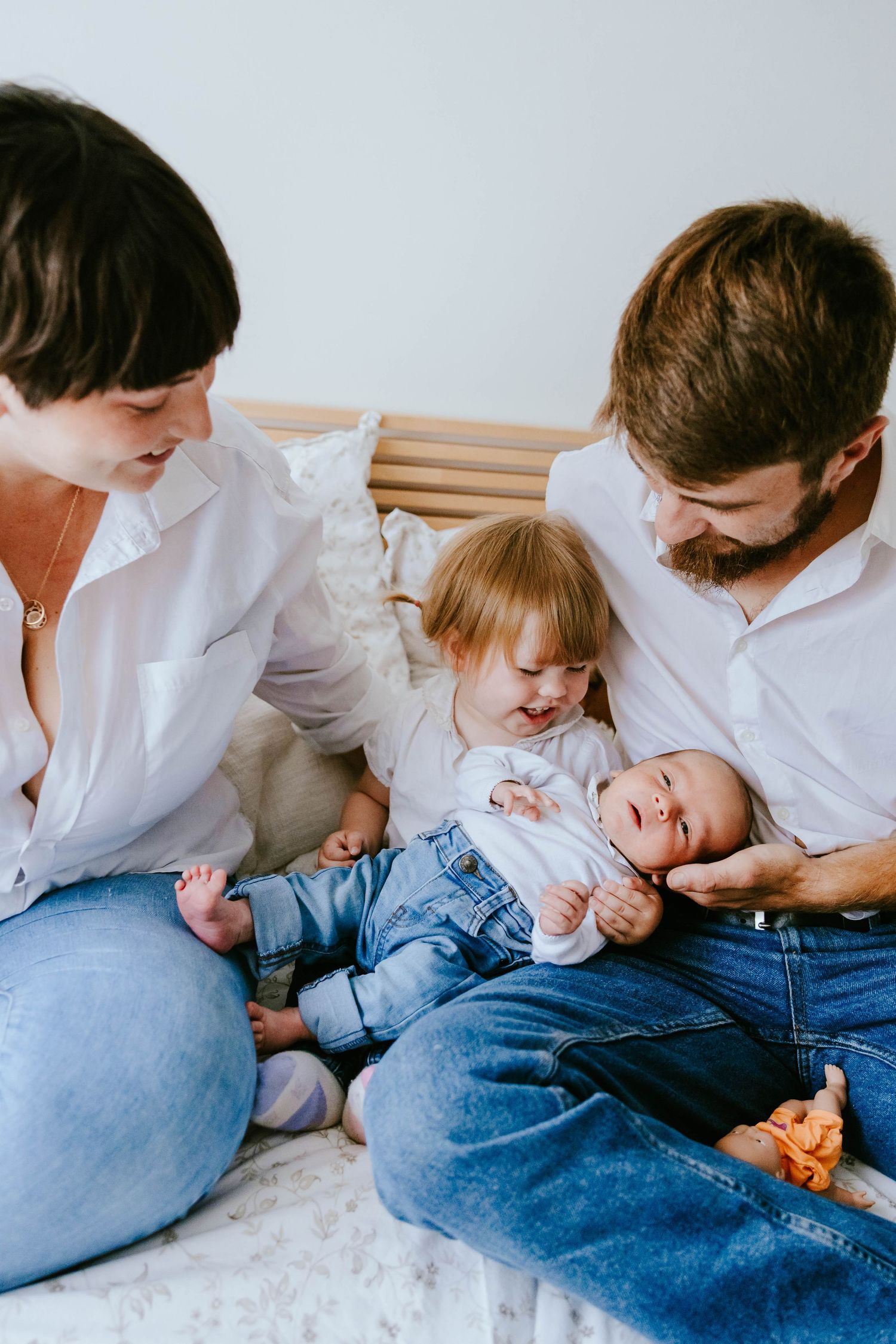 Two parents in white shirts lovingly watch their toddler interact with a newborn baby on a bed.