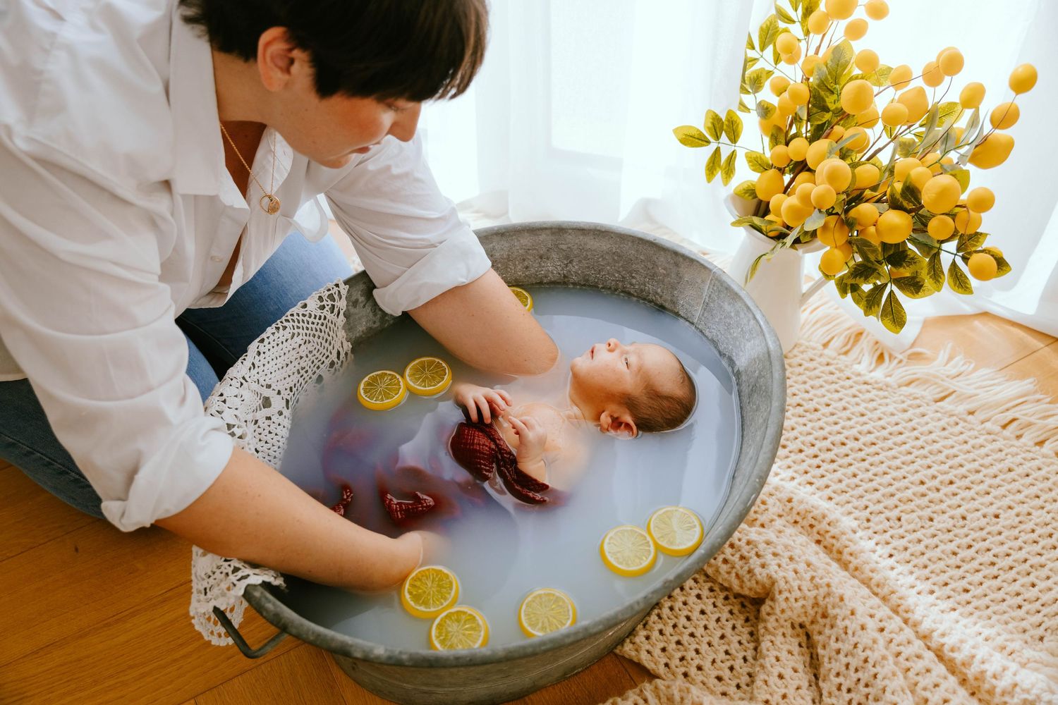Mother gently bathing newborn baby in decorative tub with lemon slices and floral water.