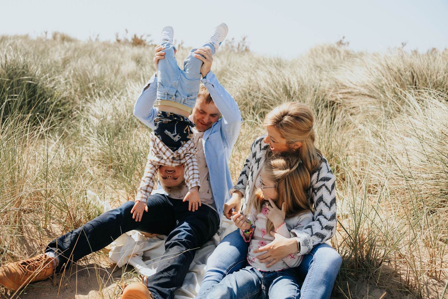 A joyful family of four plays in sandy beach dunes, with dad lifting a child upside down outdoors.