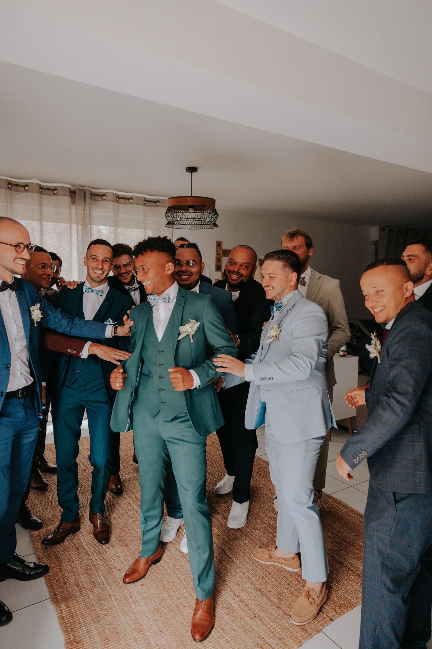 A groom in a green suit celebrates with groomsmen in blue suits, dancing joyfully indoors before a wedding ceremony.